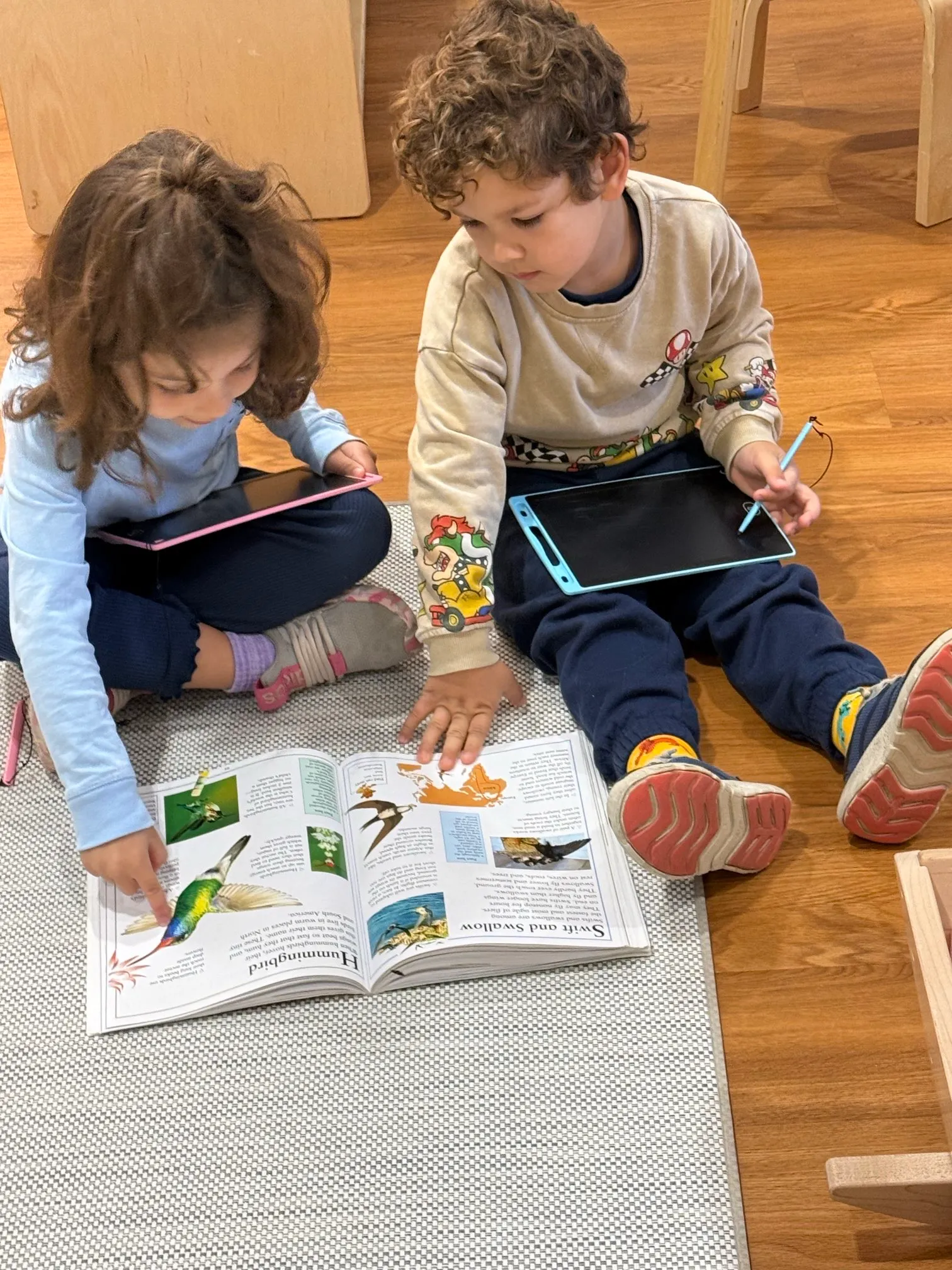 Two young children sitting on the floor with tablets, looking at a book about hummingbirds and swifts.