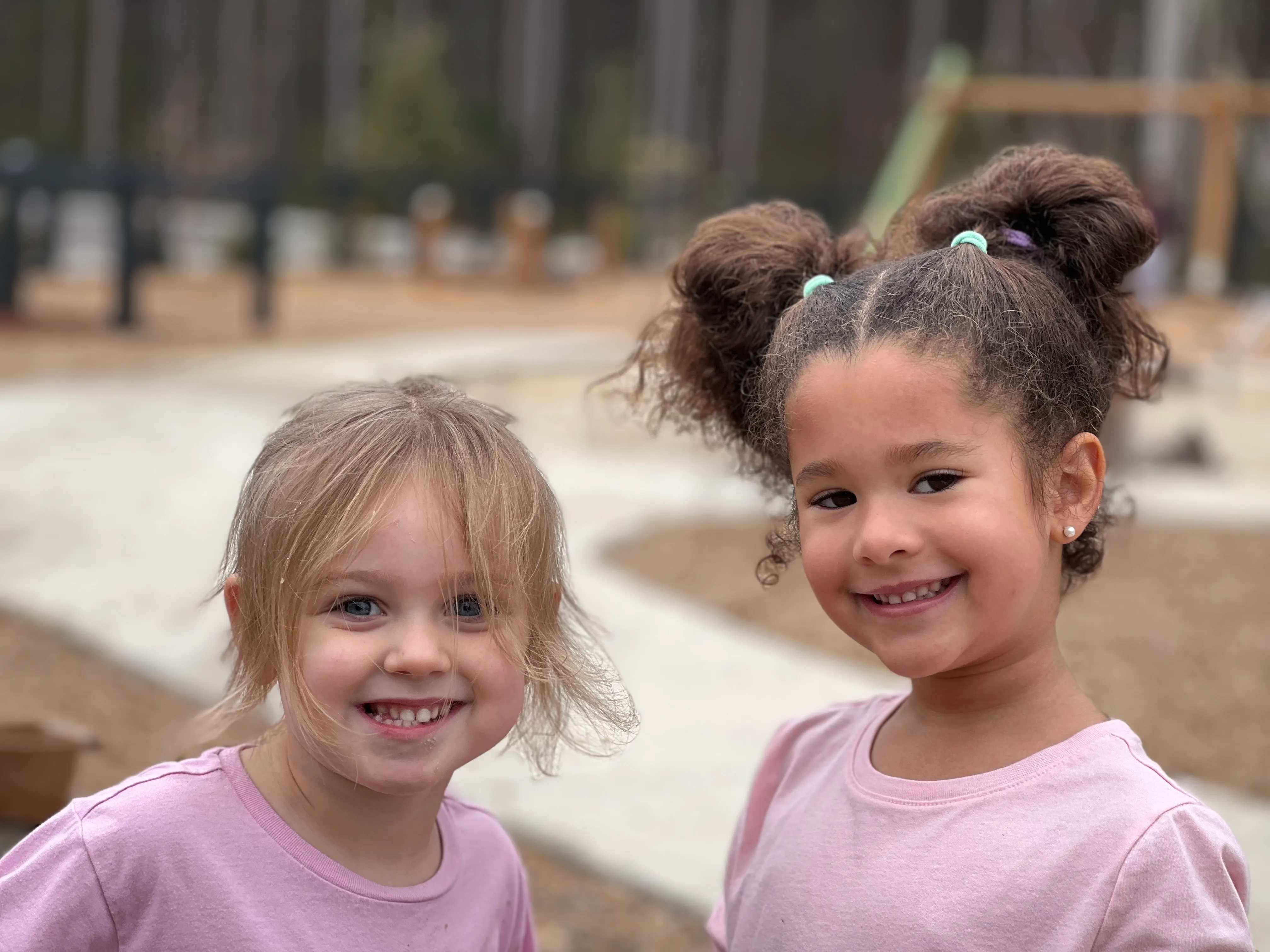 Two young girls smiling outdoors, both wearing light pink shirts, with a blurred playground background.