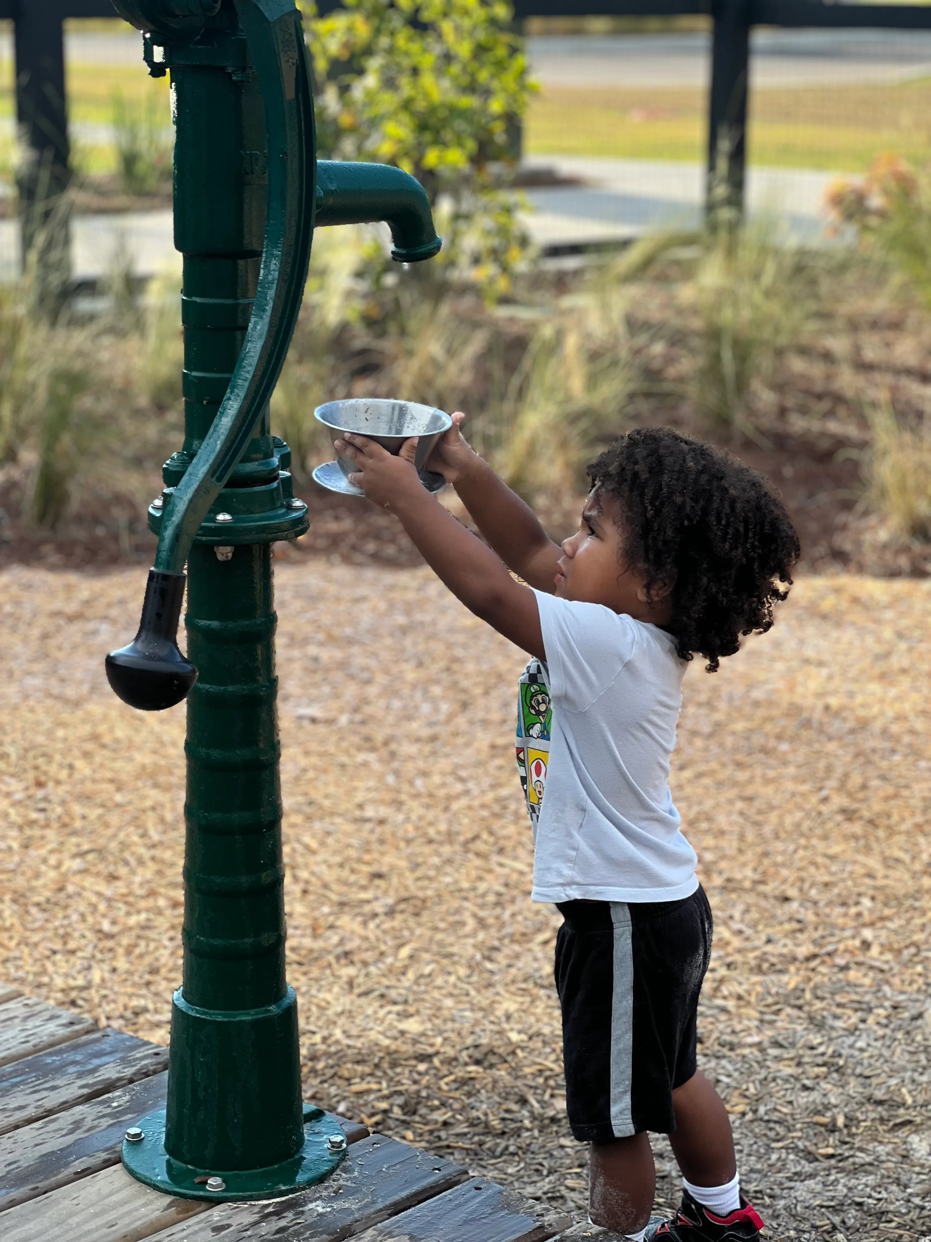 Young child holding a metal cup up to an old-fashioned green water pump outdoors.