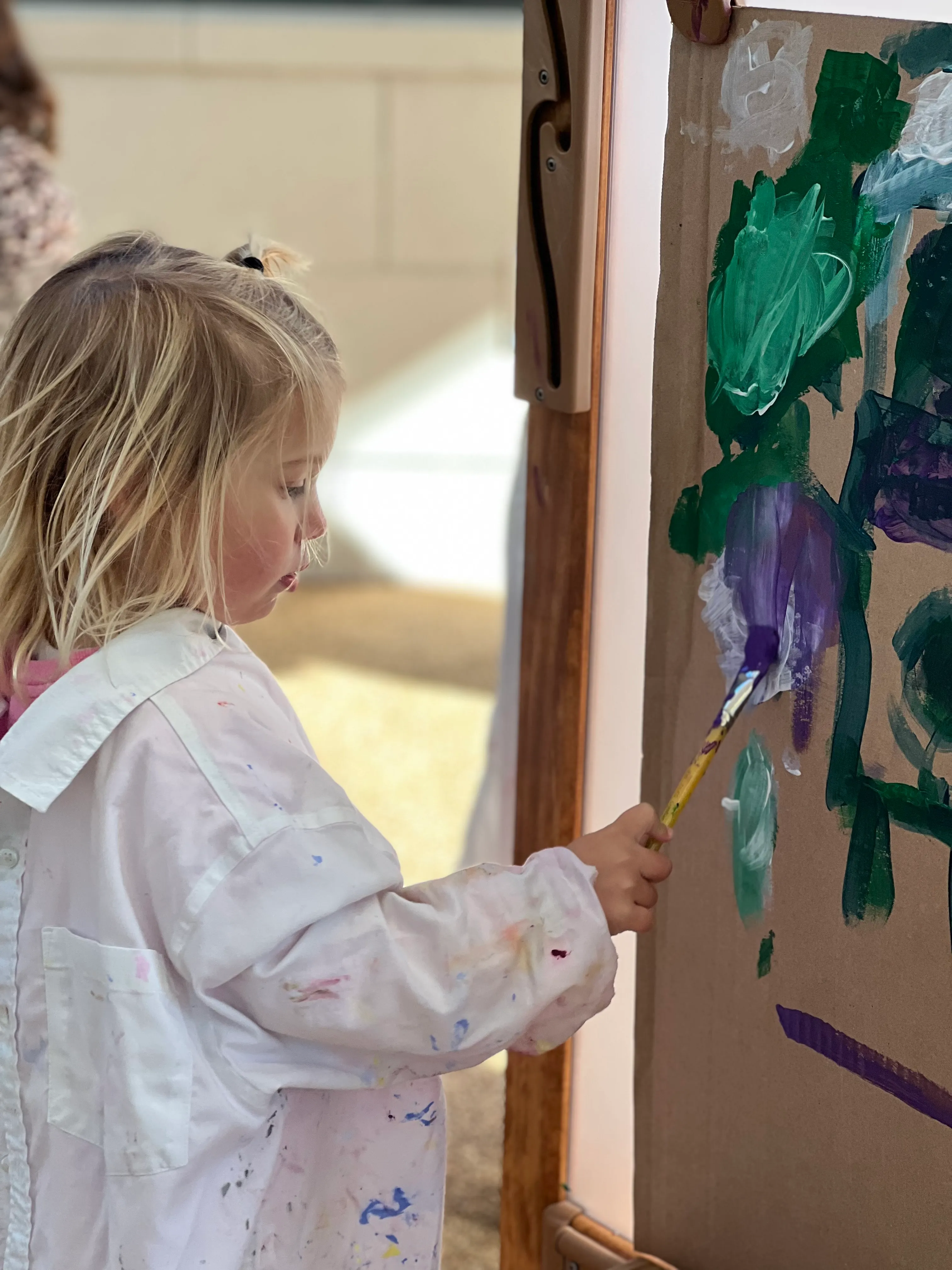 Young child wearing a paint-stained smock painting with a brush on a vertical paper-covered easel.