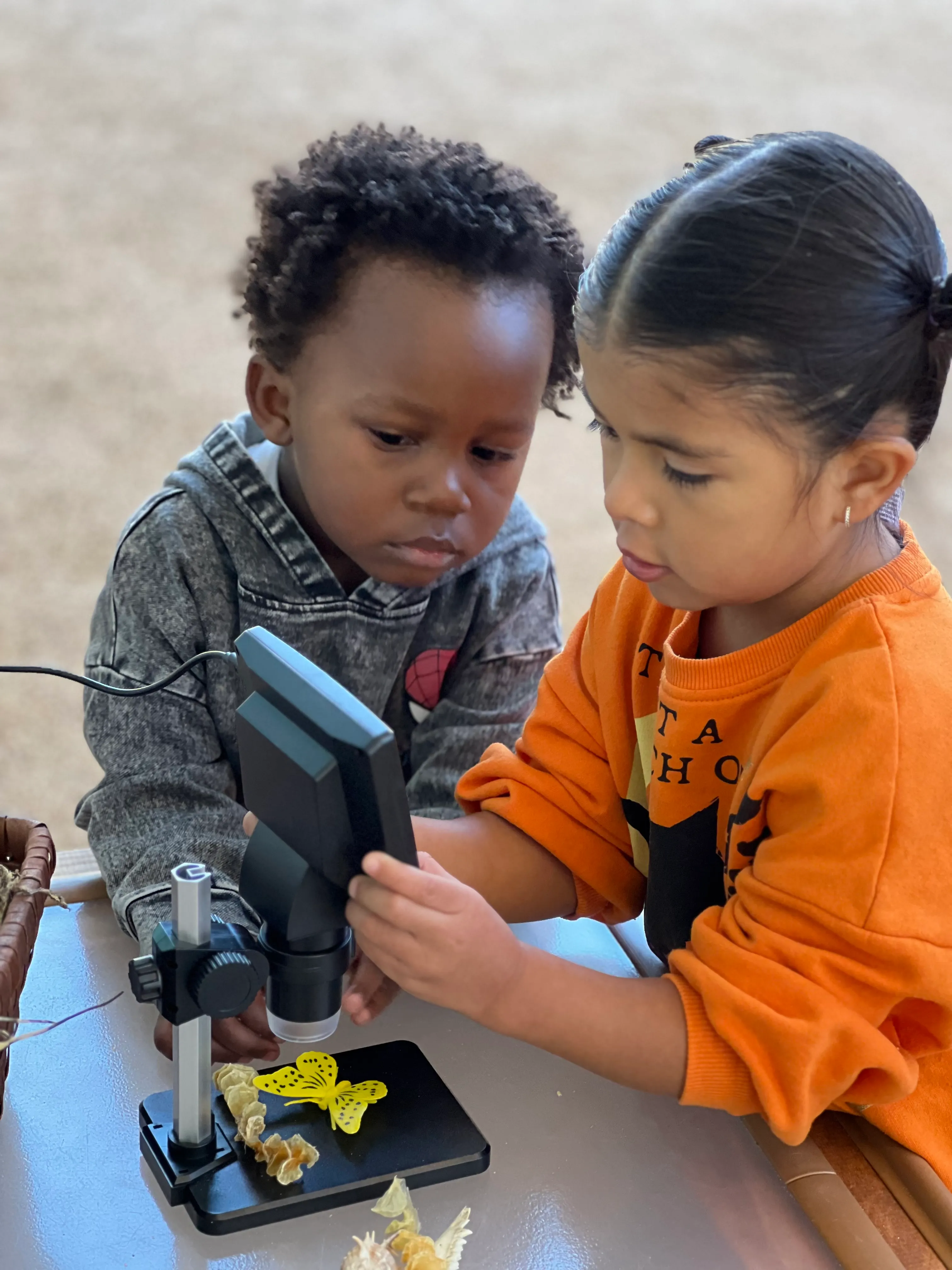 Two children examining a yellow paper butterfly under a digital microscope on a table.