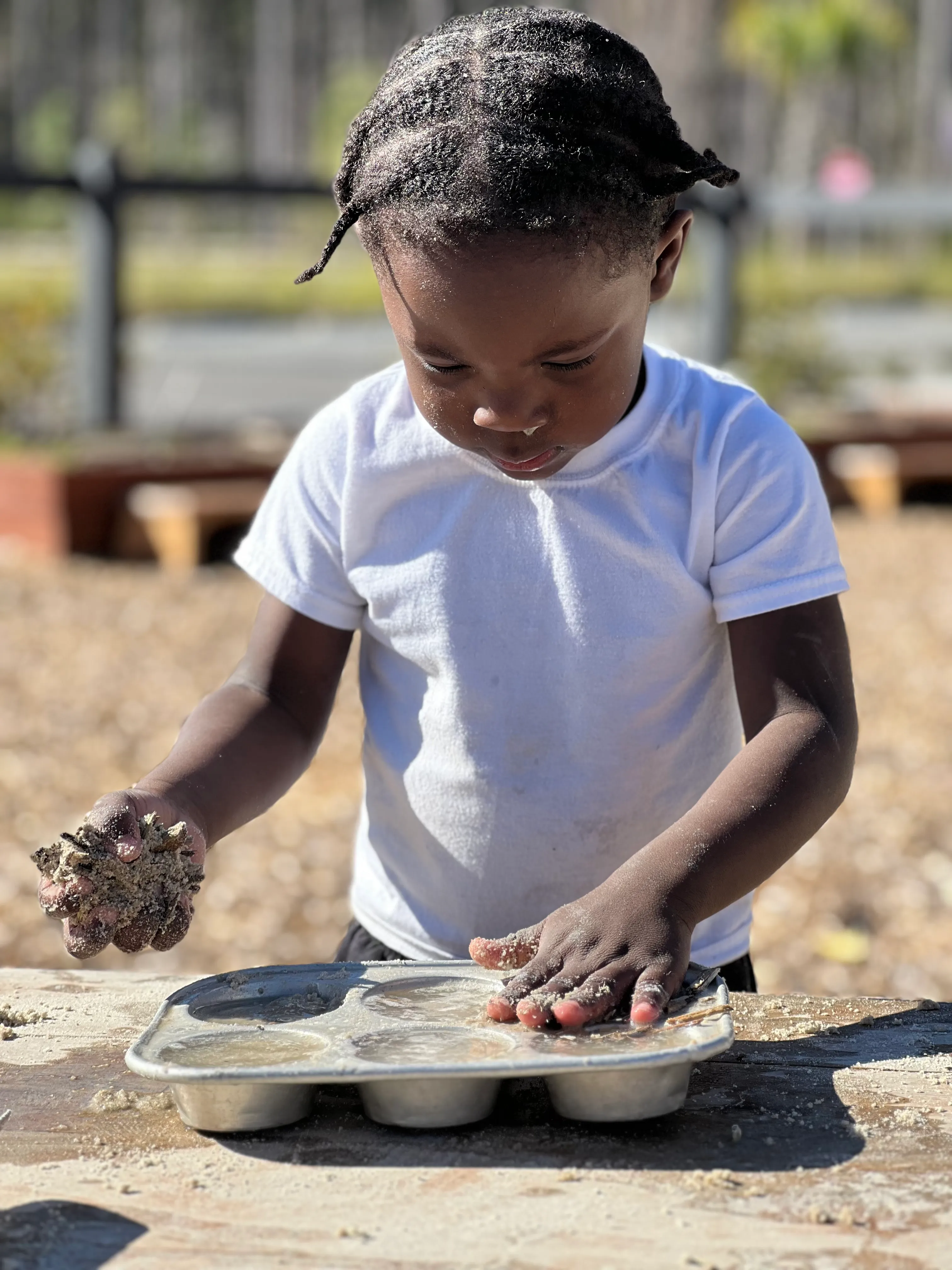 Young child playing outdoors with sand, pressing it into a metal muffin tin.
