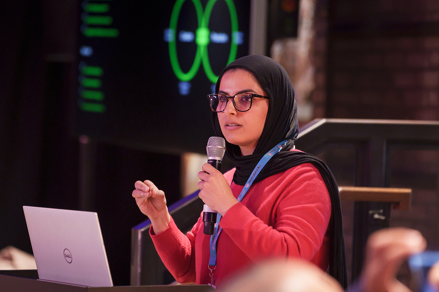 MLOps speaker in a headscarf presenting with a microphone beside a laptop and projected diagram