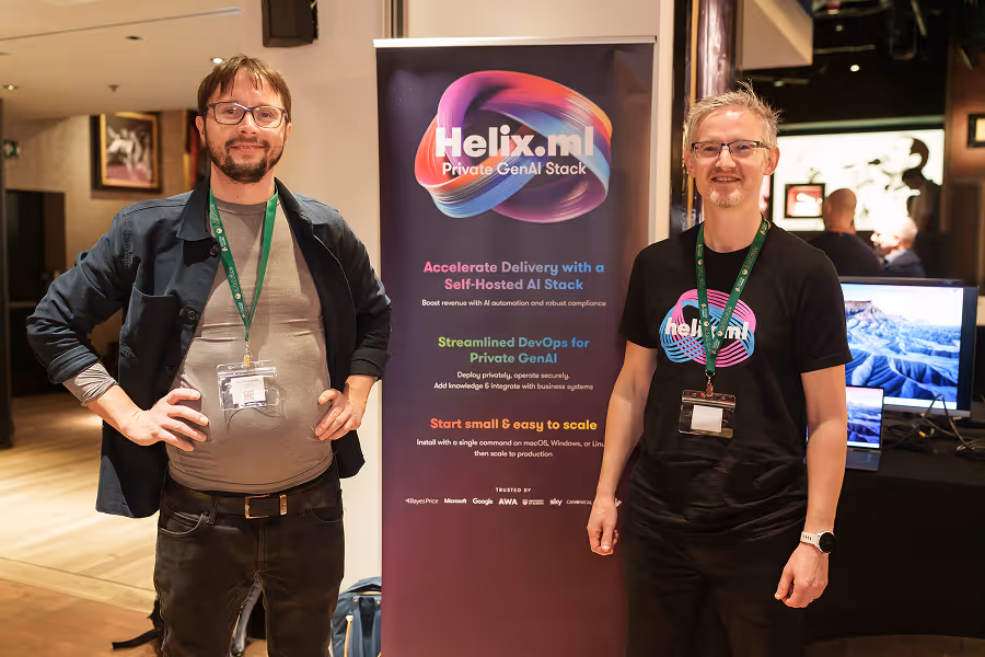 Two men with glasses and lanyards standing confidently beside Helix.M AI stack banner at MLOps conference booth