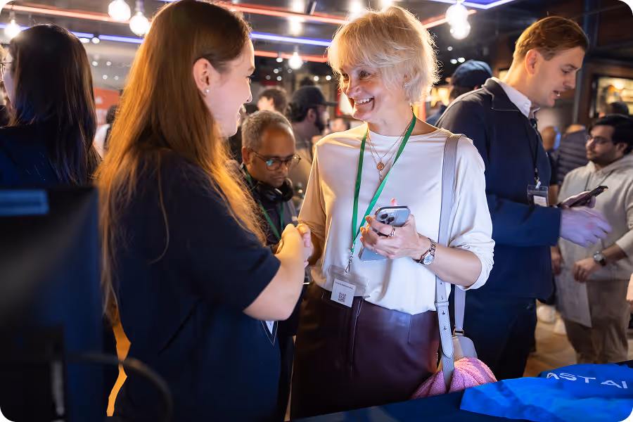 Two women with lanyards chatting at an MLOps event booth near Astai booth banner