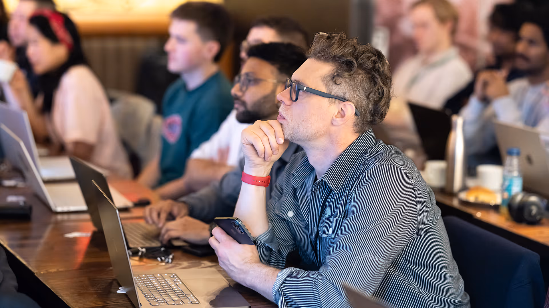 Man with curly hair and glasses thoughtfully resting chin on hand at a conference table with laptops