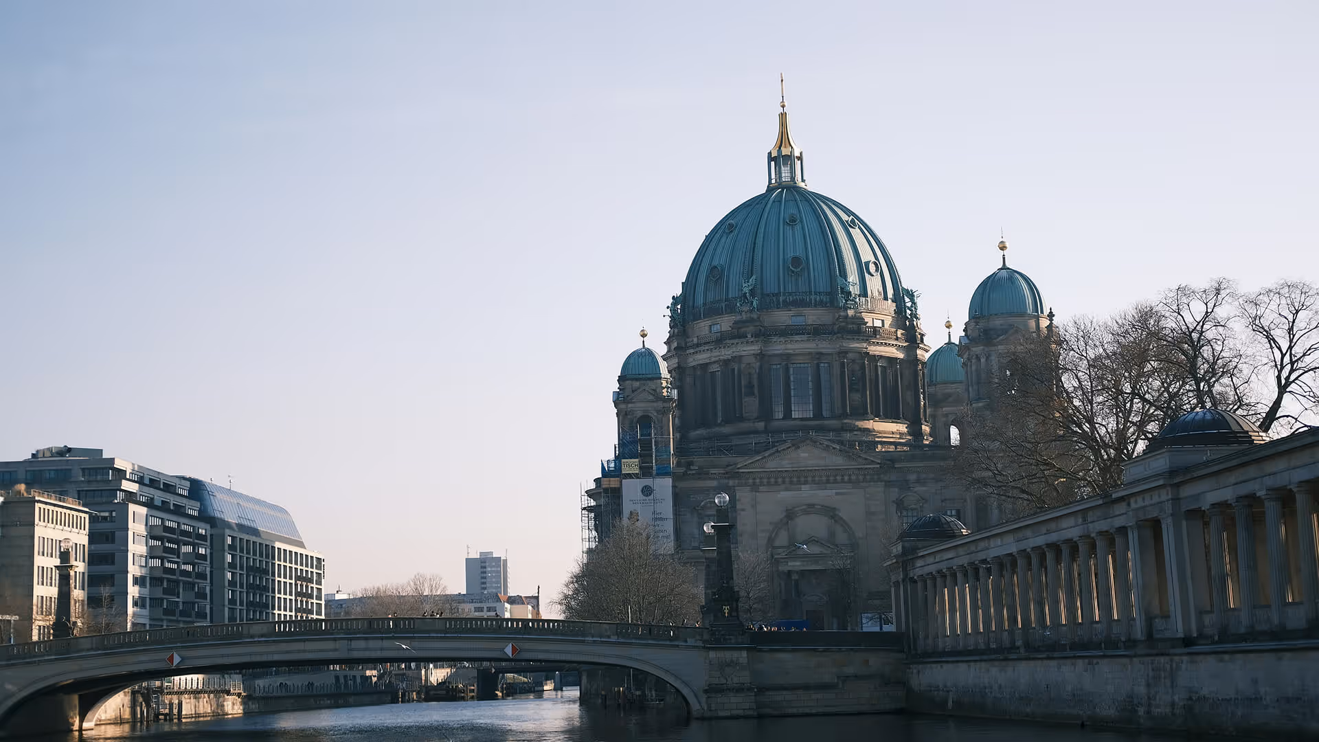 Berlin Cathedral with green dome and gold spire beside river, bridge, and modern buildings