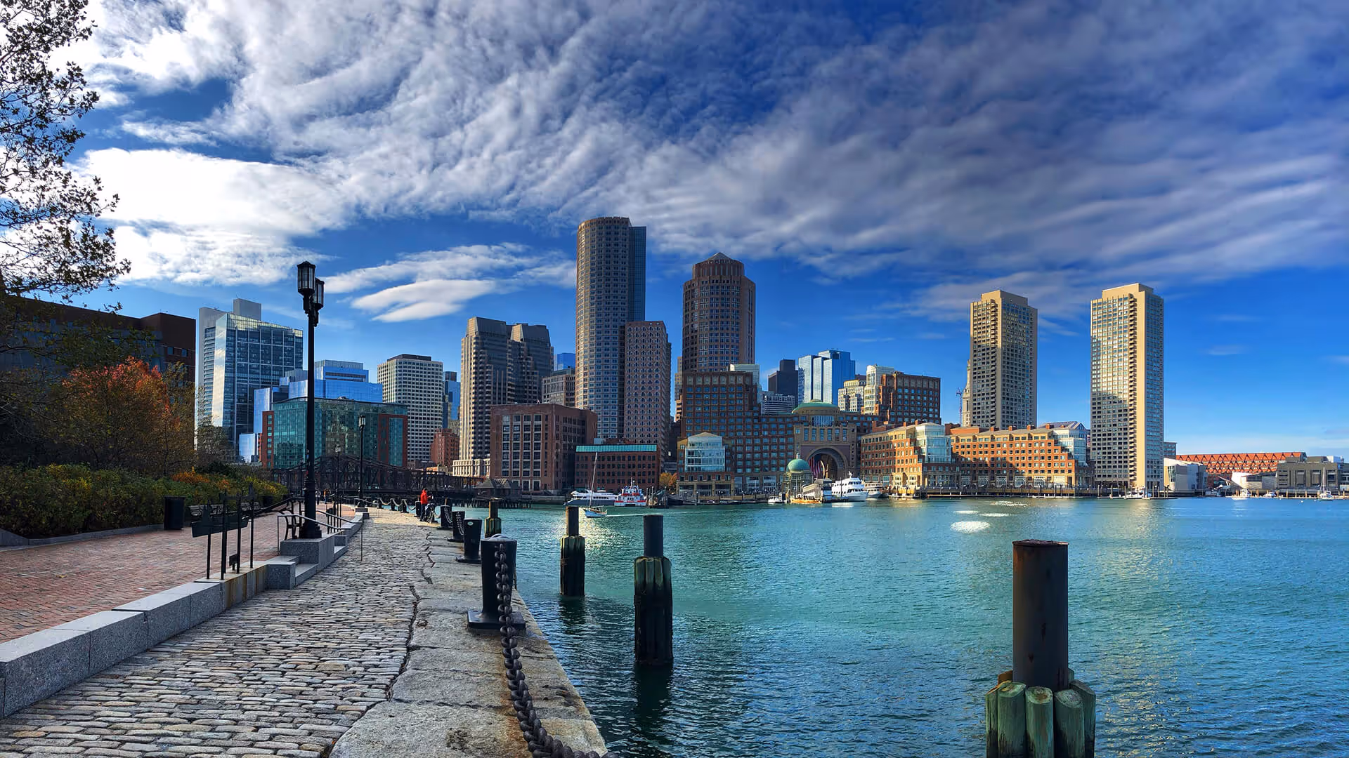 Boston skyline with tall skyscrapers, autumn trees, brick walkways, and harbor boats under cloudy skies