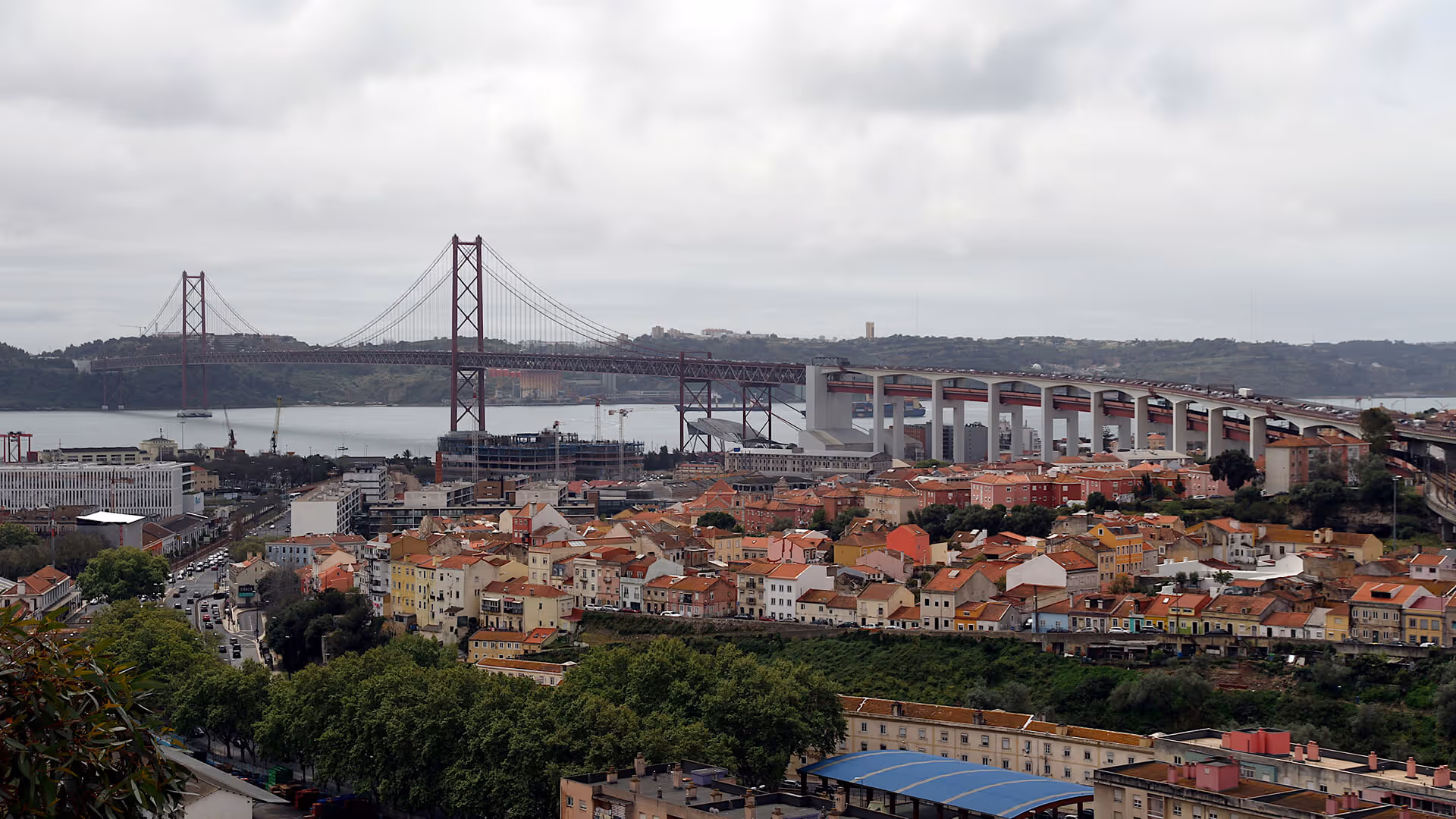 Lisbon waterfront with iconic 25 de Abril red suspension bridge, terracotta rooftops, cranes, and Tagus River
