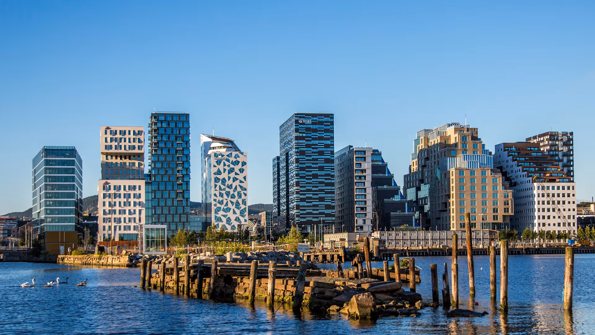 Oslo's modern angular high-rise buildings reflecting in fjord with wooden pier pilings at sunset