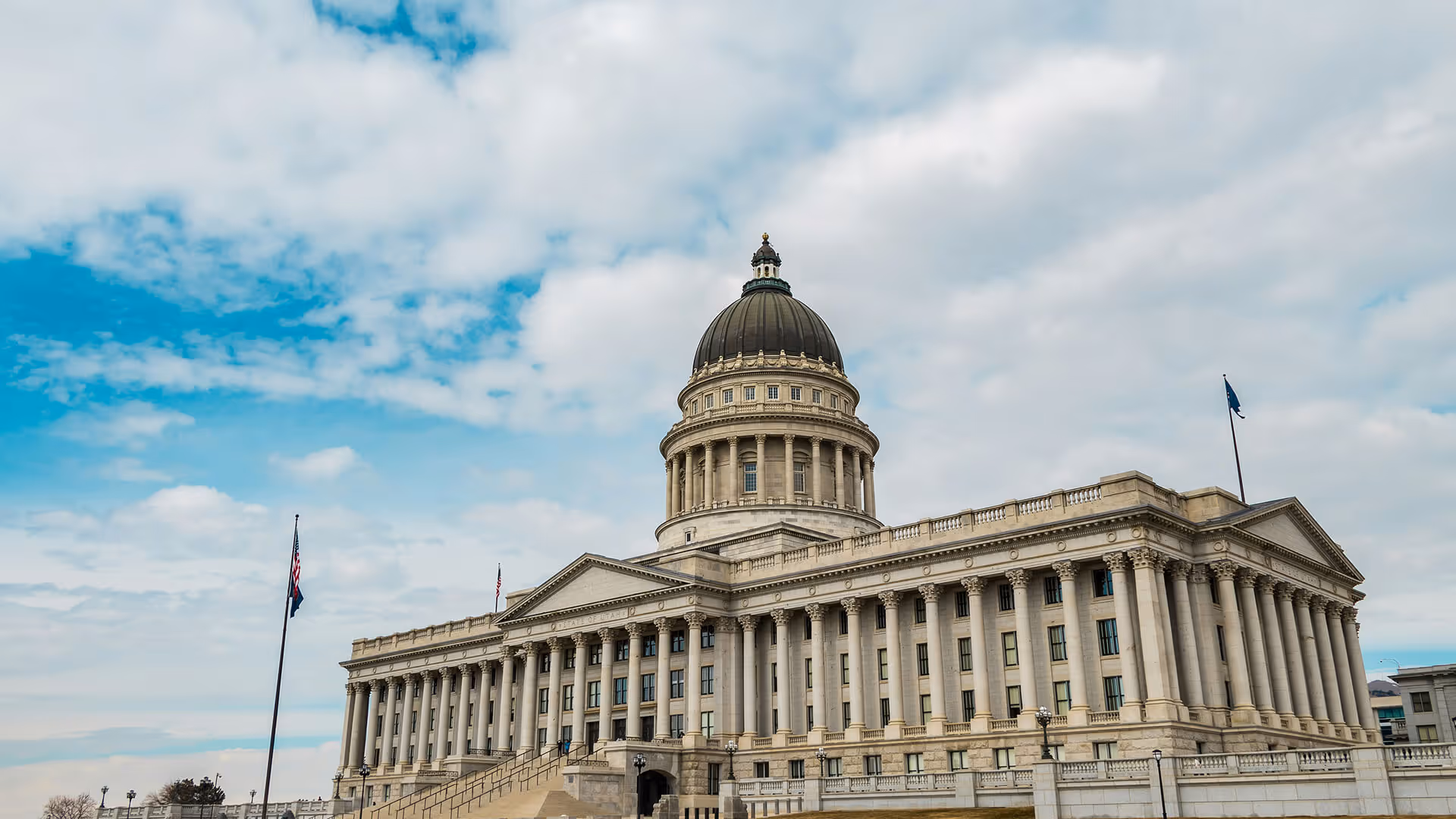 Utah State Capitol building with neoclassical dome, columns, and flags against blue sky with clouds