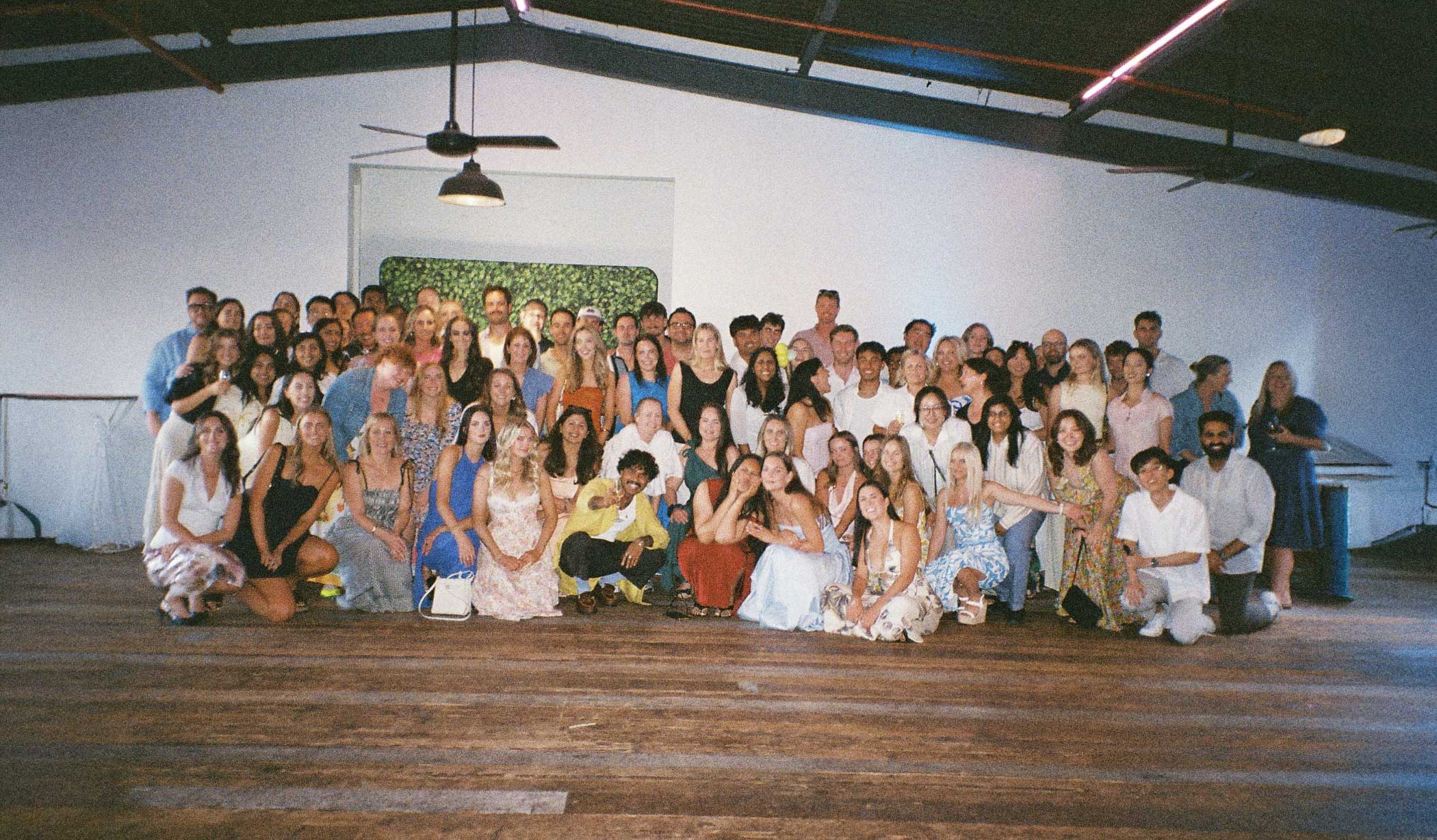 MBM's Auckland office team posing together indoors on wooden floor under ceiling fans and lights.