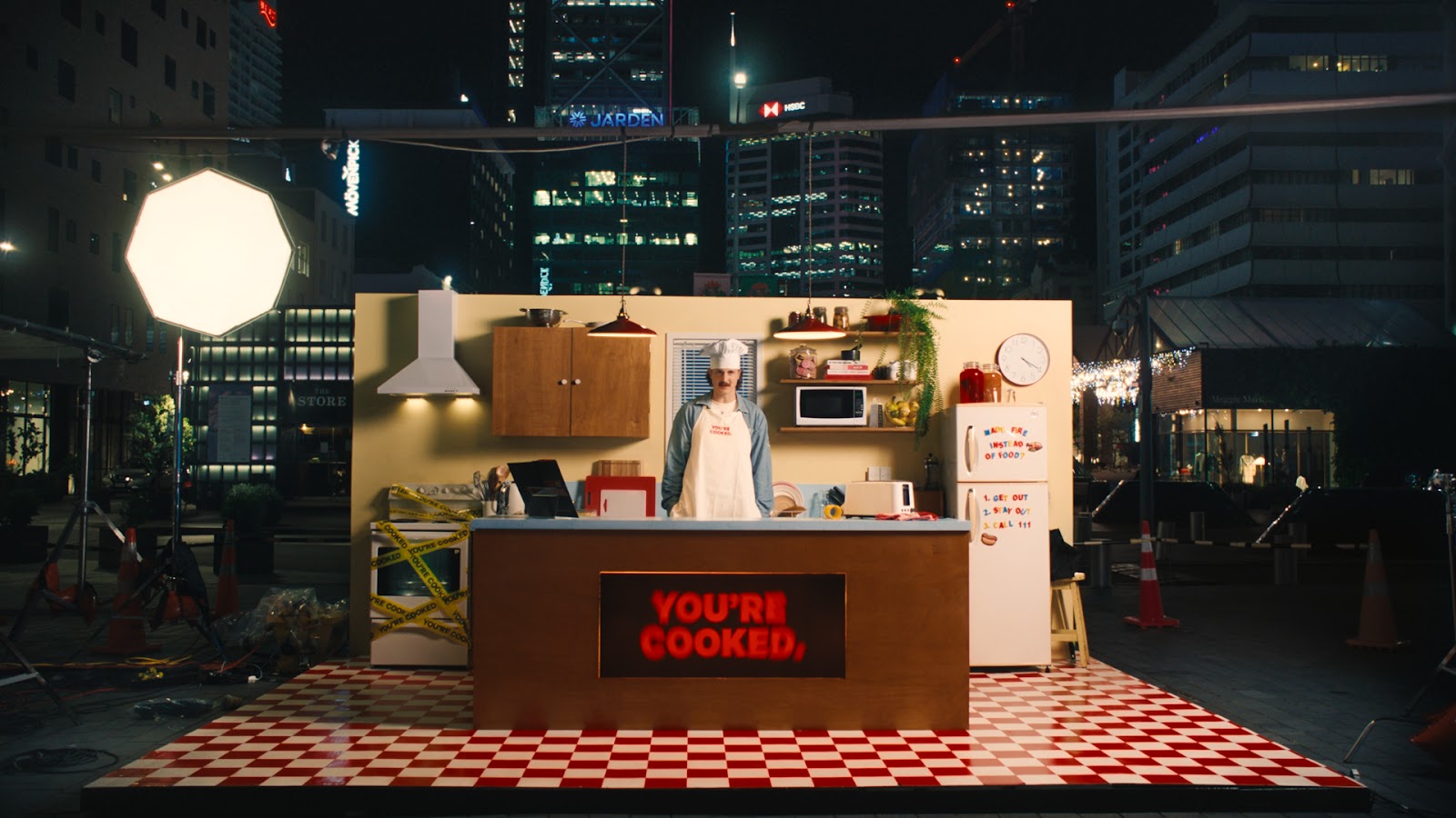 Chef standing behind a kitchen island in an outdoor nighttime city set with a lit sign reading 'YOU'RE COOKED,' and a refrigerator with safety instructions.