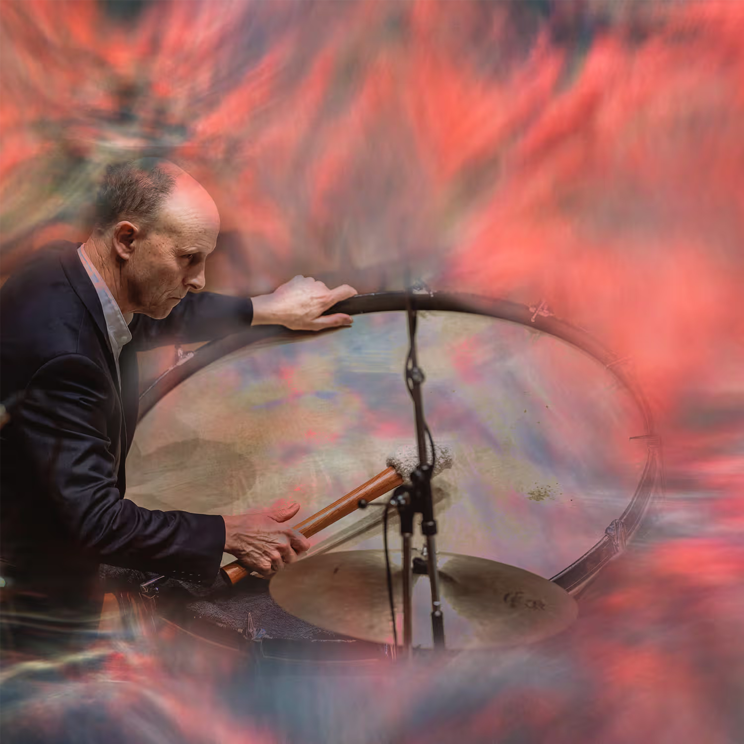Gary Wain of the Tasmanian Symphony Orchestra playing a large drum with drumsticks, set against a vibrant, abstract background of swirling red, pink, and blue clouds, blending musical performance with artistic expression.