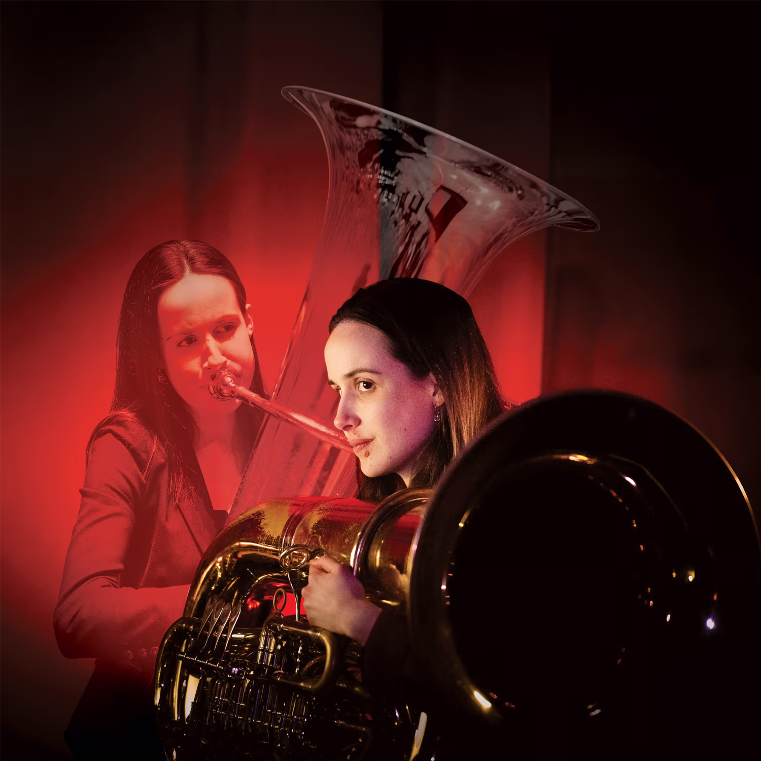 Tuba player Rachel Kelly performing under a red spotlight in a dimly lit setting. She is shown in profile with long hair and dark clothing, playing a tuba with its large bell and intricate tubing prominently visible. A superimposed image of Rachel playing 