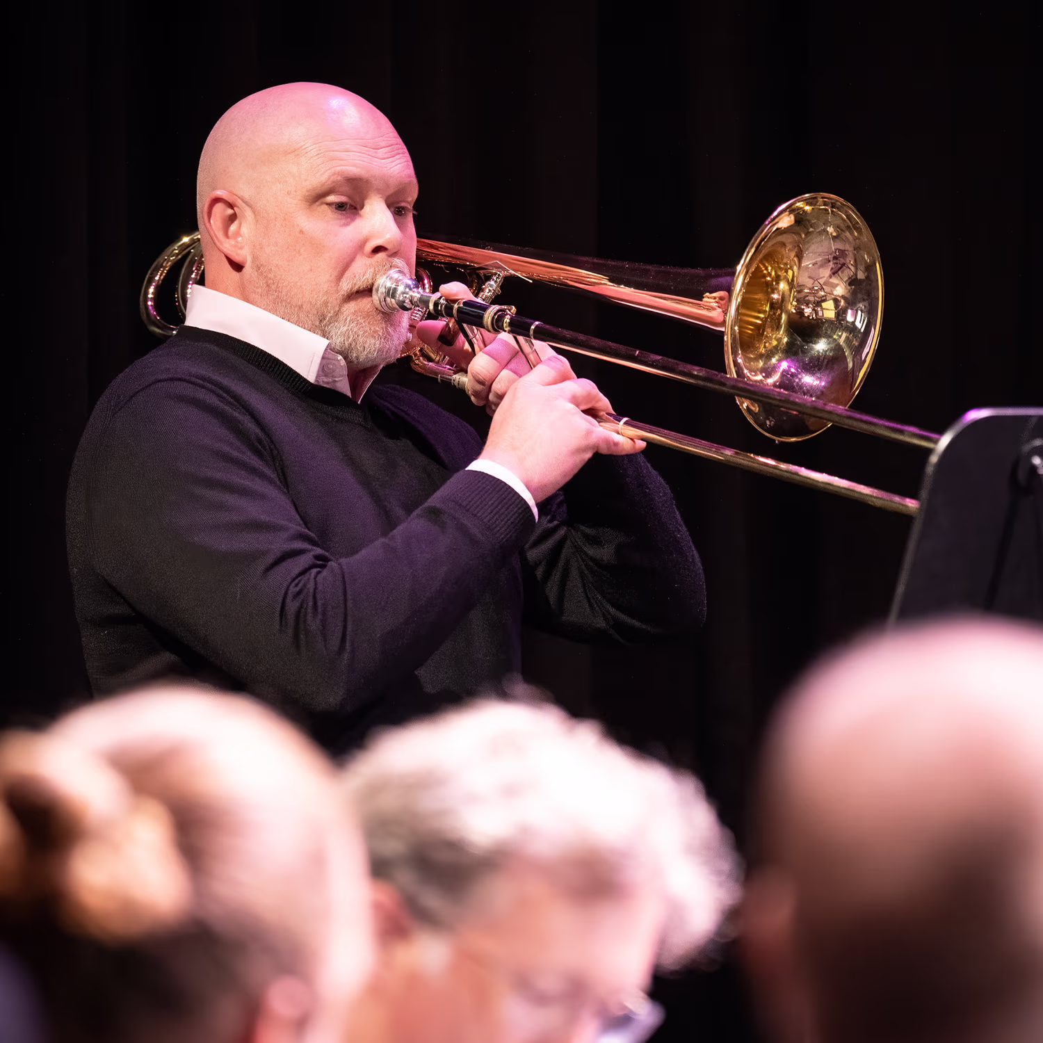 Close-up of a musician playing trombone on stage, audience blurred in foreground under warm lights