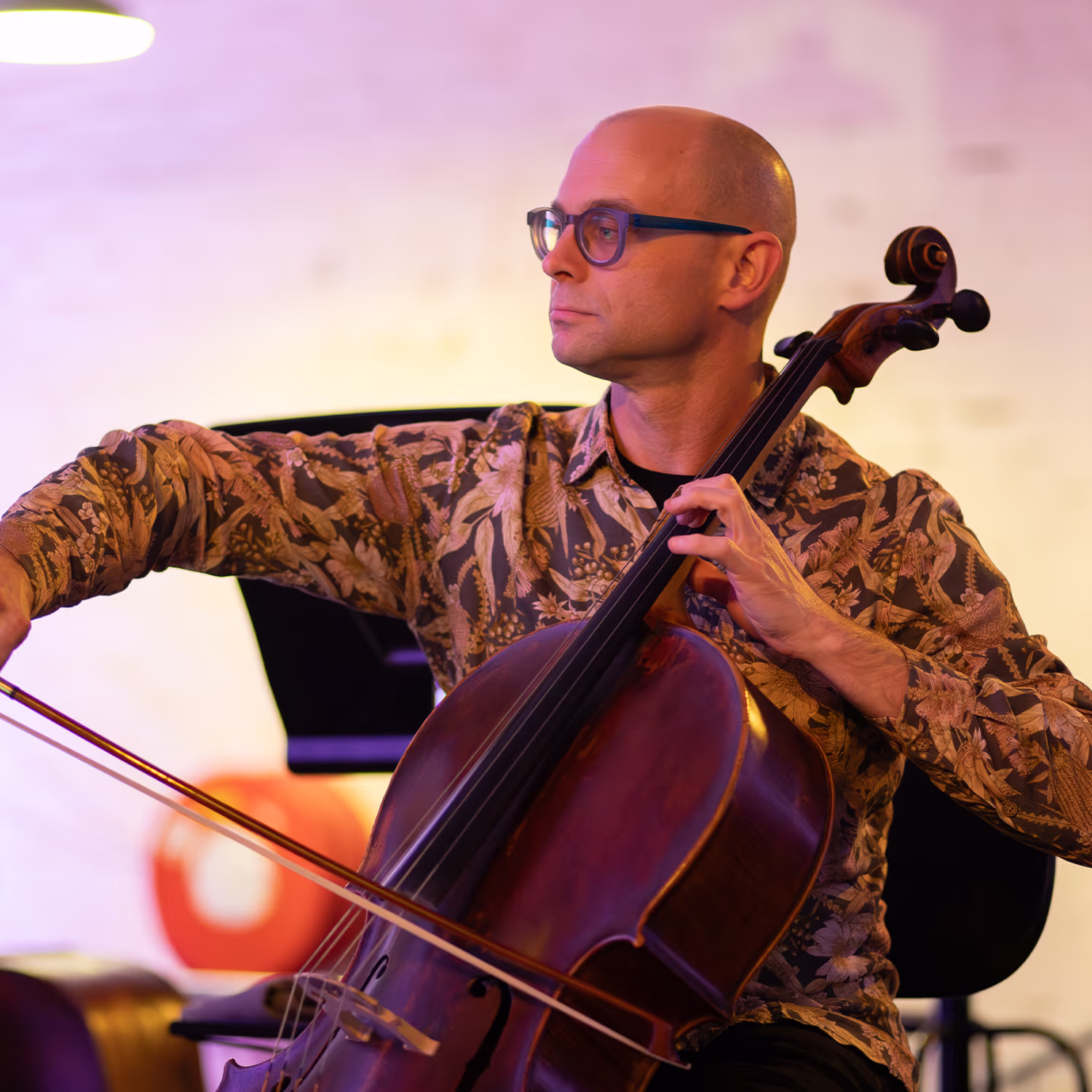 Close-up cellist with glasses playing cello on stage, warm lighting and blurred audience foreground