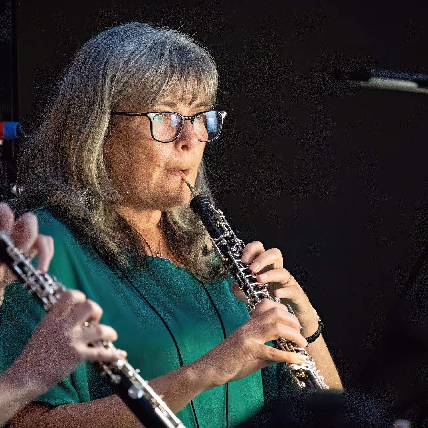Close-up of musician with glasses playing oboe on stage, another oboe visible, dark backdrop