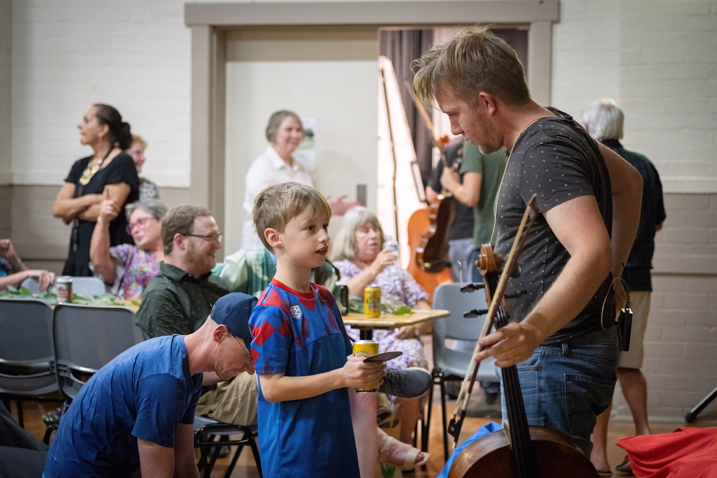 A blonde man carrying a cello stops to talk with a young boy in a community hall.