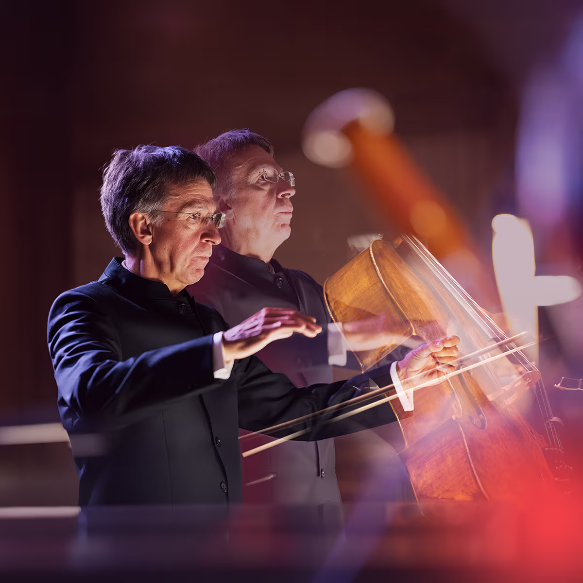 A man in a black suit gestures with his hands in the foreground while a cellist plays behind him.