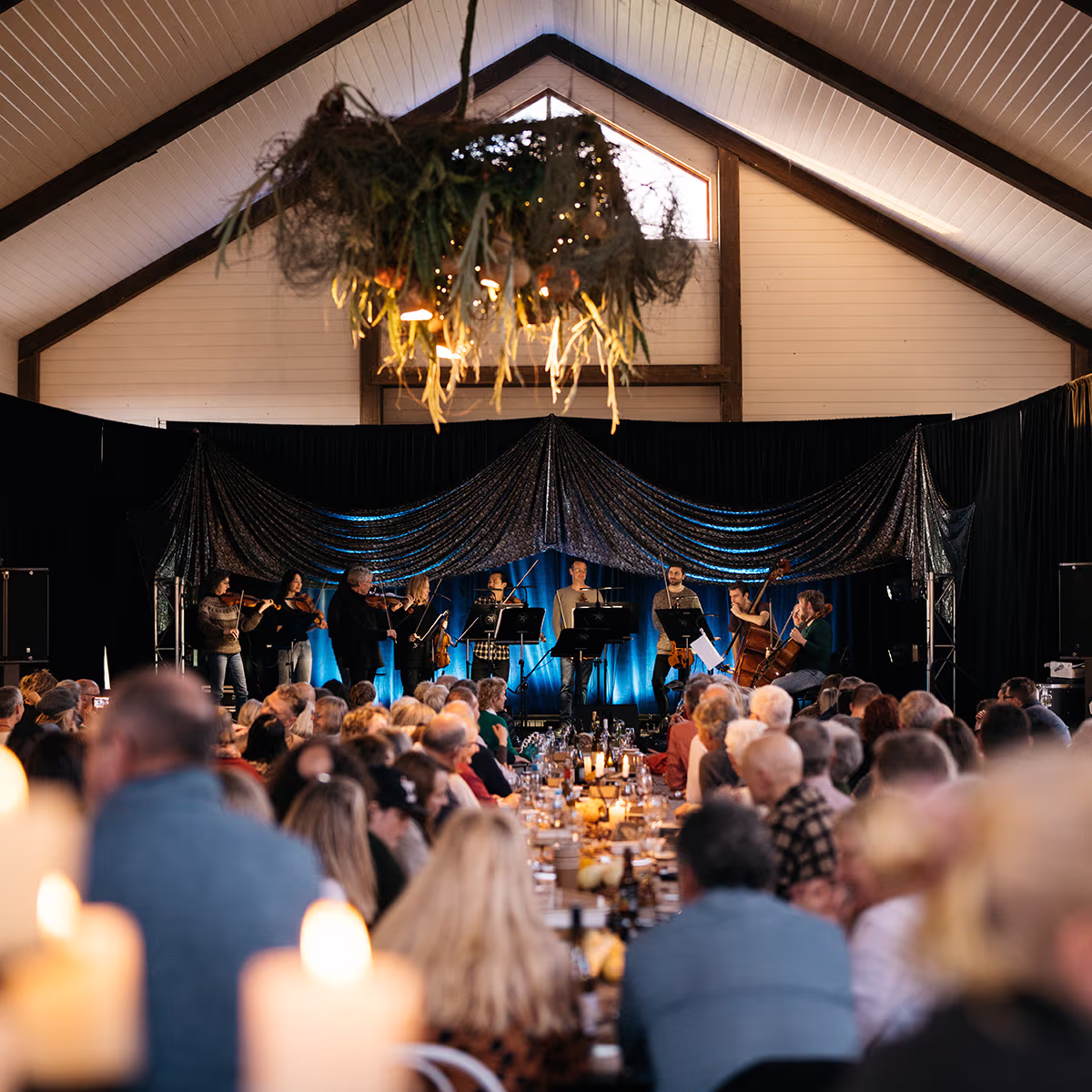 An audience sits at long farm tables eating food and watching a live performance.
