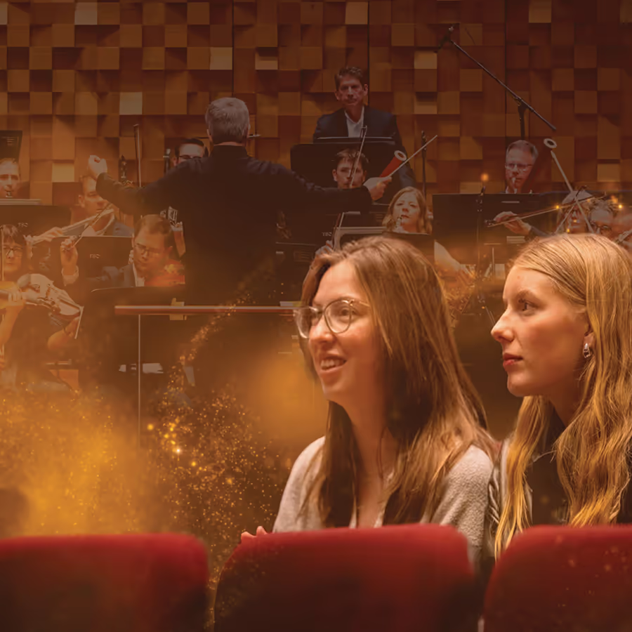 Two audience members watch an orchestra during an Open Rehearsal as a conductor leads musicians onstage behind them. Warm concert hall lighting and red seats create an intimate view of the rehearsal experience.