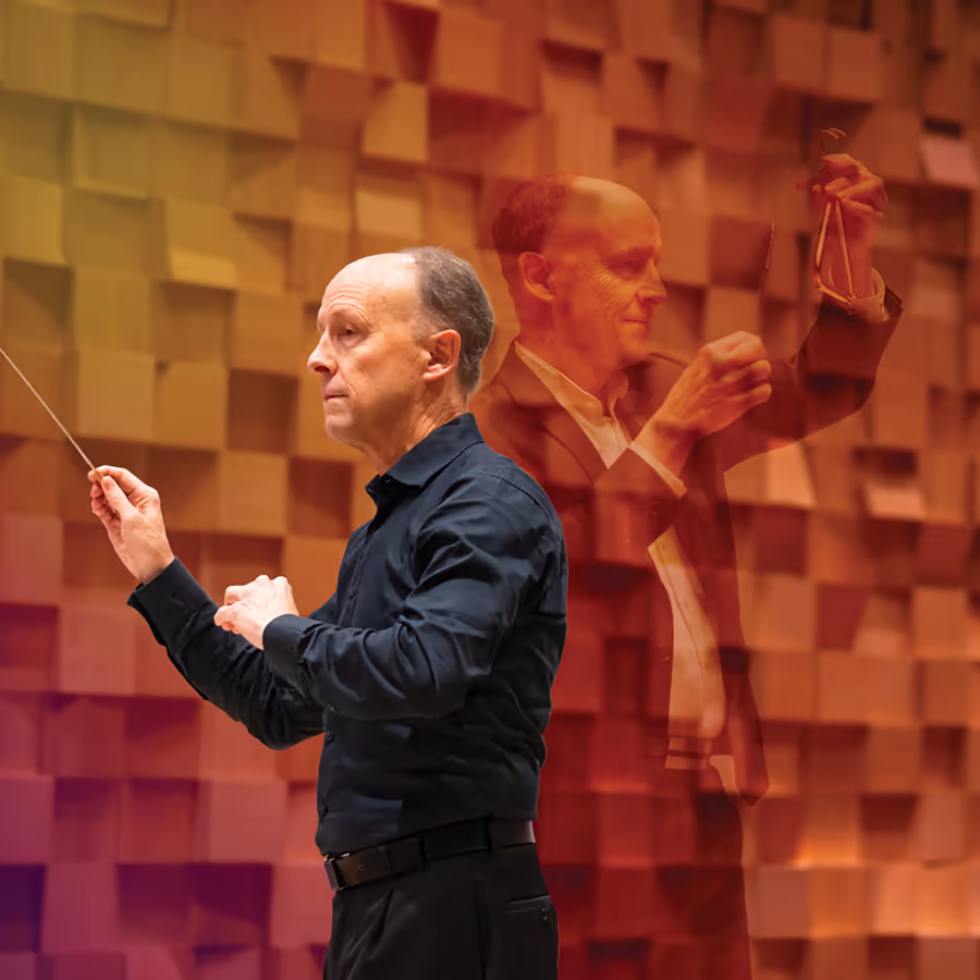 Conductor Gary Wain holds a baton while leading a conducting session against a warm orange acoustic wall.