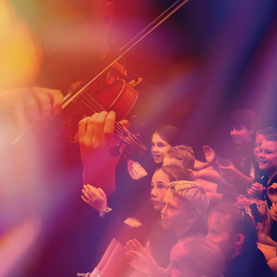 A violinist plays up close while a group of children watch and clap from their seats. Warm orange pink and purple stage lighting creates an energetic atmosphere during the live performance.