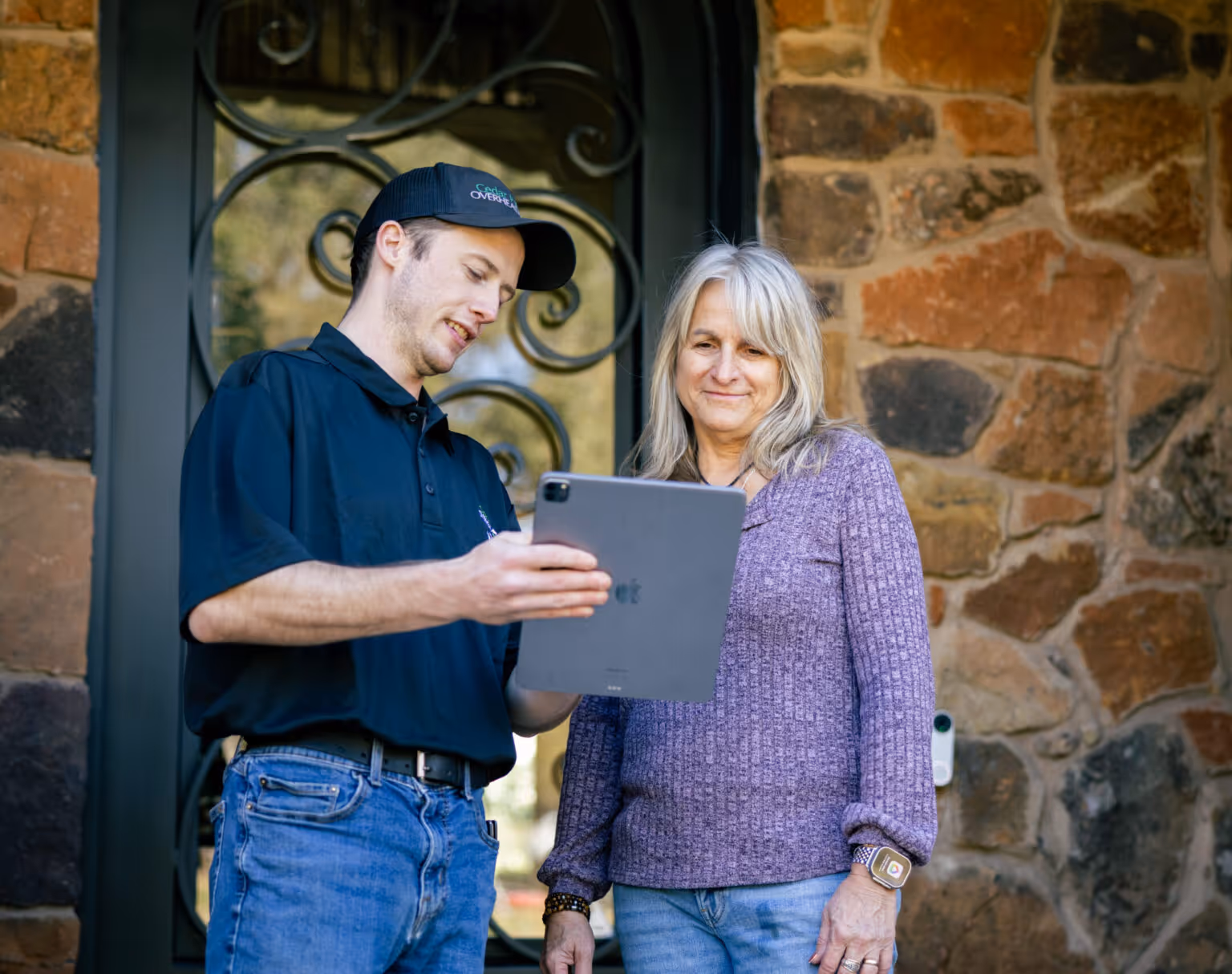 Man in a black polo and cap showing a tablet to a smiling woman in a purple sweater standing outside a stone building.