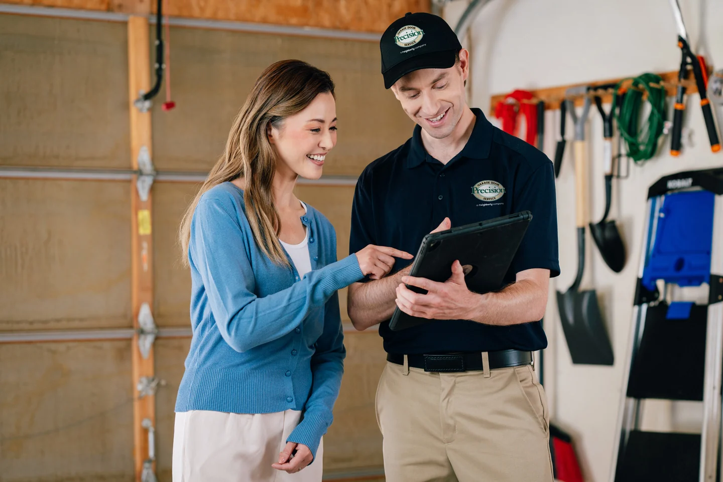 A technician in a Precision Service uniform shows a tablet to a smiling woman in a garage with tools hanging on the wall.