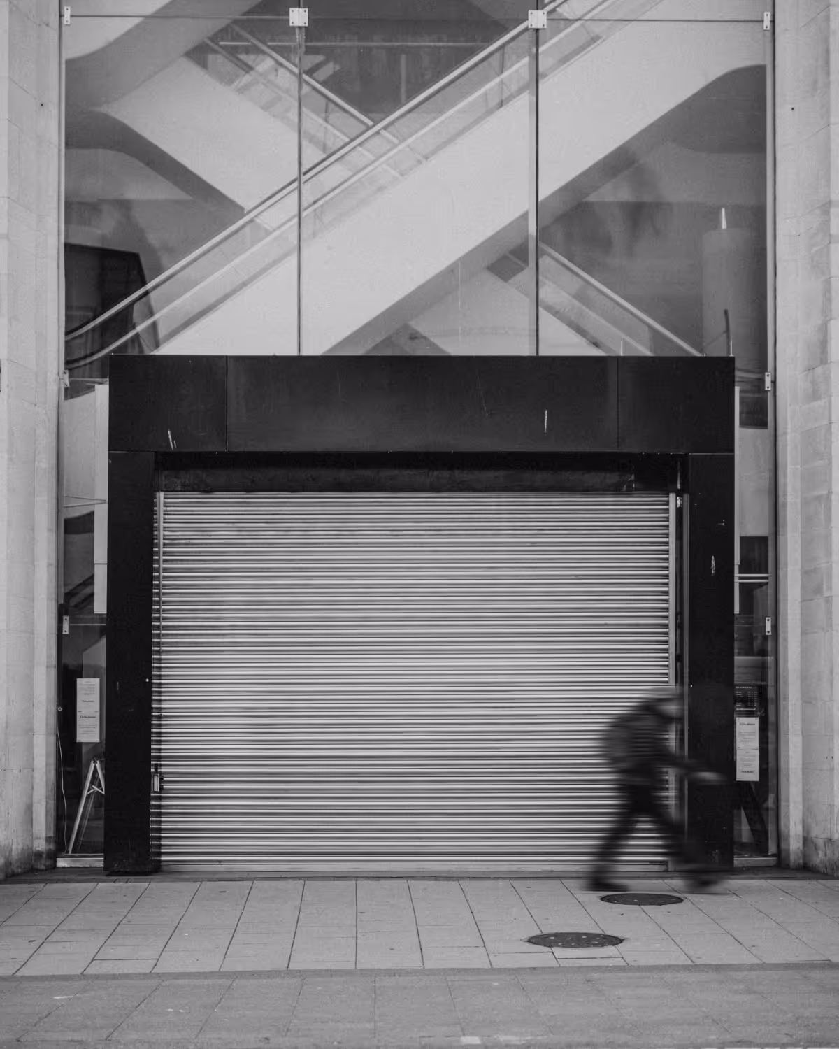Black and white photo of a closed metal shutter with a blurred person riding a scooter in front and glass escalators visible behind.
