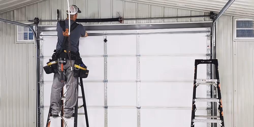 Technician on ladder repairing or installing a white metal garage door inside a metal building.