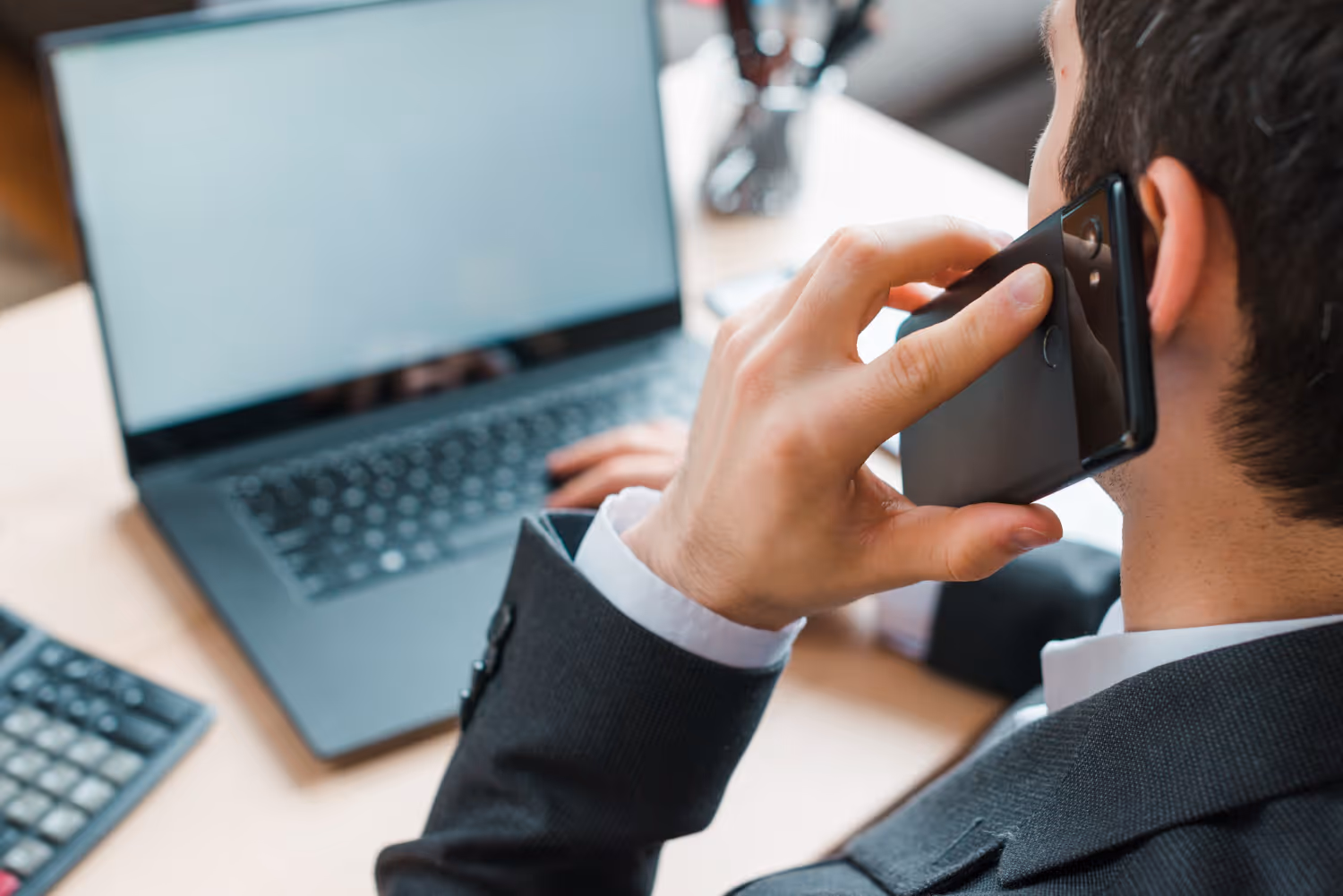 Man in suit talking on a smartphone while working on a laptop at a desk.