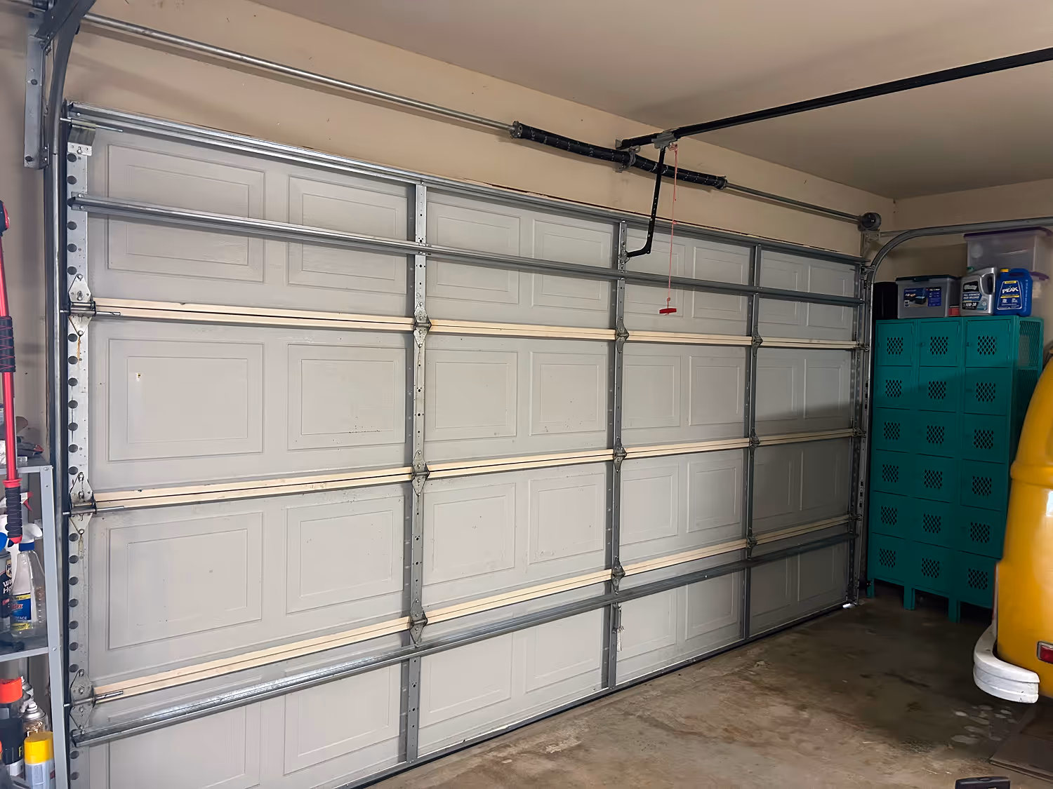 Closed metal garage door in a garage with storage cabinets and various containers on the right.