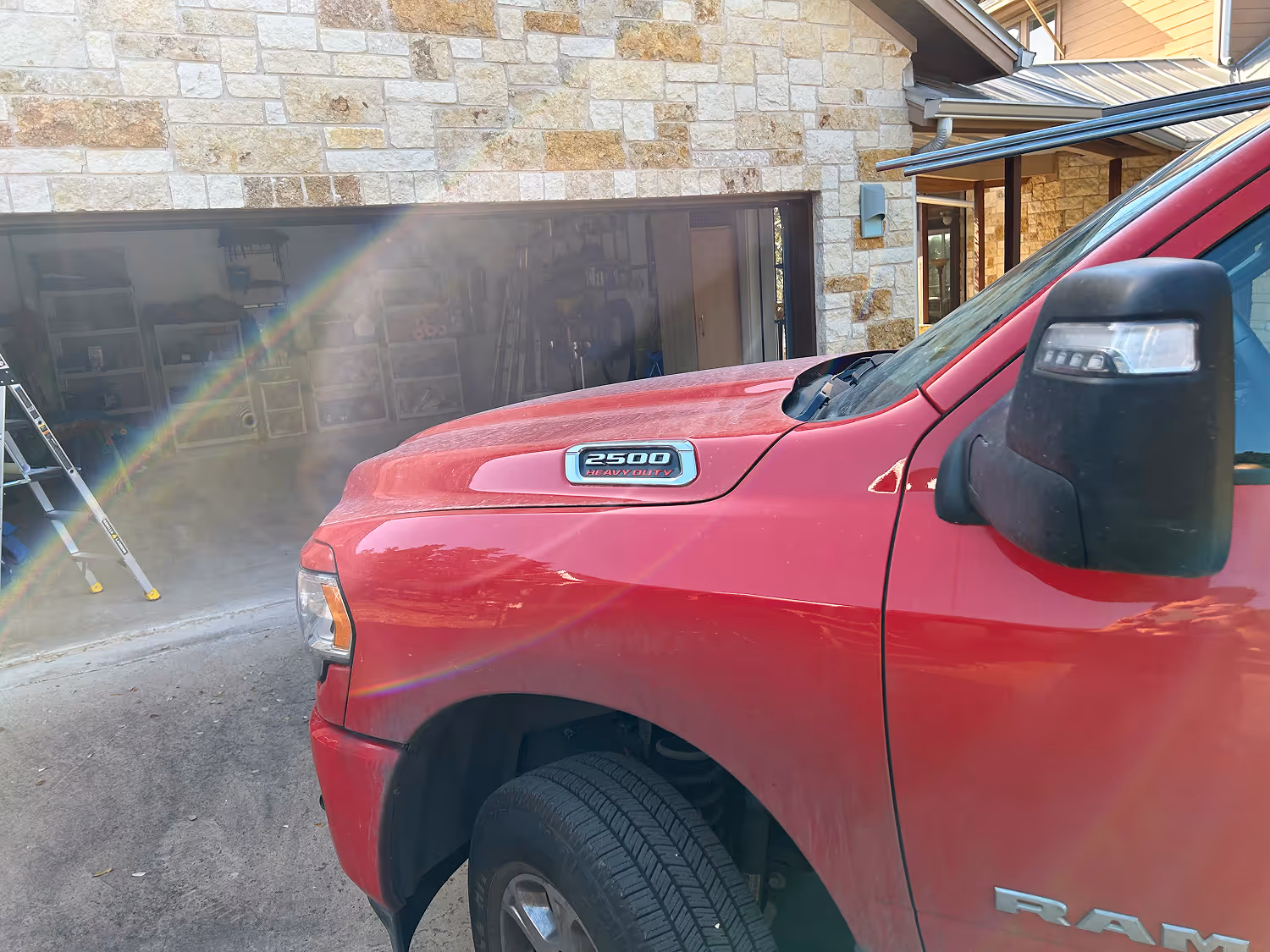 Front side view of a red Ram 2500 heavy duty truck parked in front of a stone garage with an open door and a ladder inside.