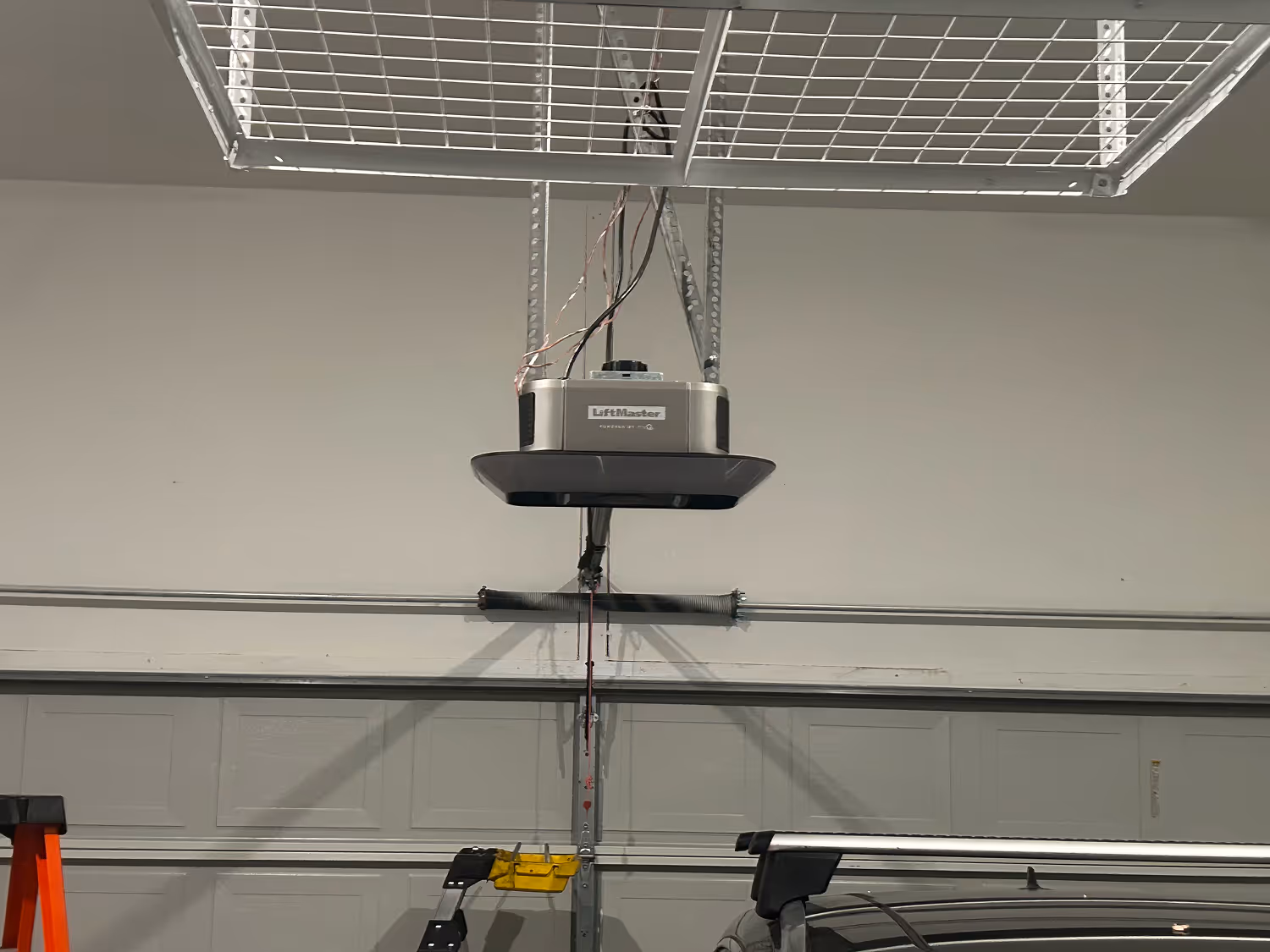 Ceiling-mounted LiftMaster garage door opener with exposed wires and partially visible ladder and car roof in a garage.