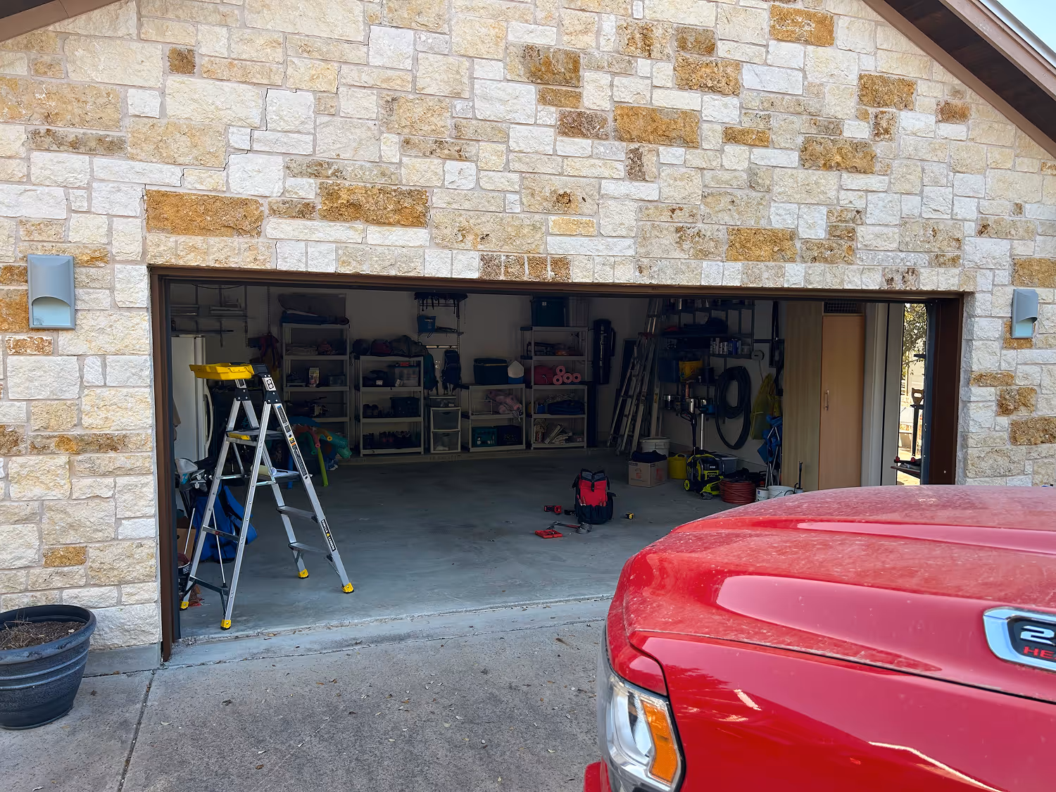 Open garage with stone exterior showing a folded silver step ladder inside and the front of a red vehicle parked outside.