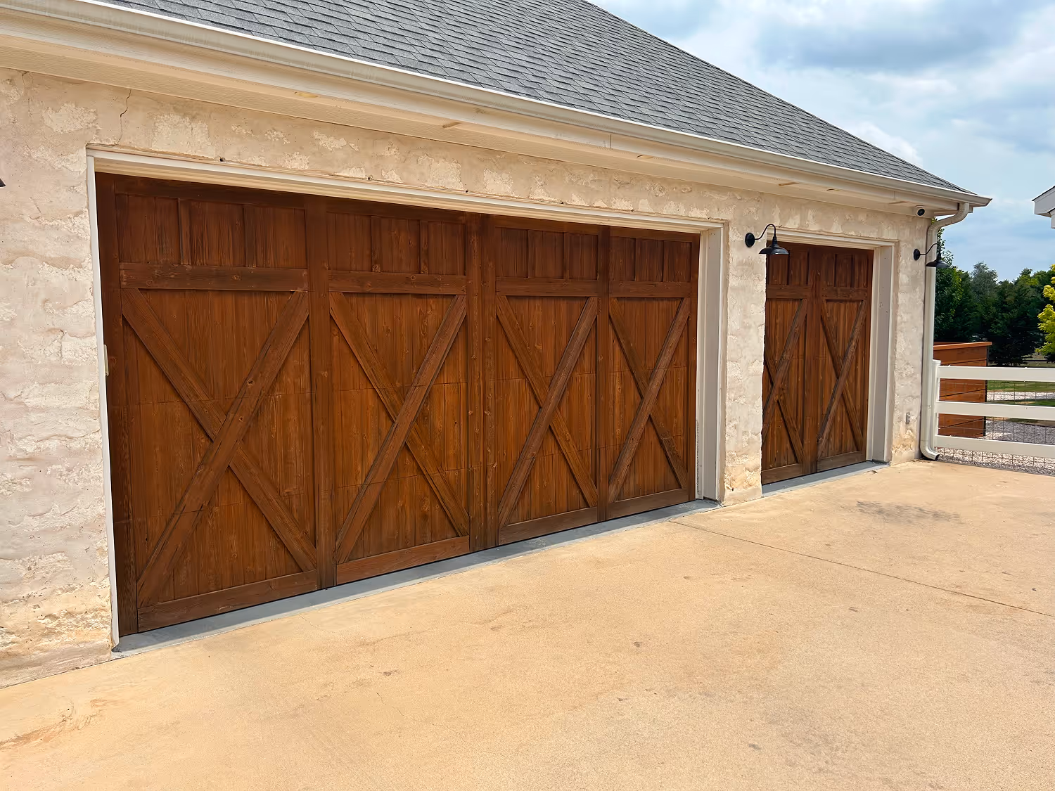 Two sets of large wooden garage doors with X-shaped paneling on a stucco building with a gray shingled roof.