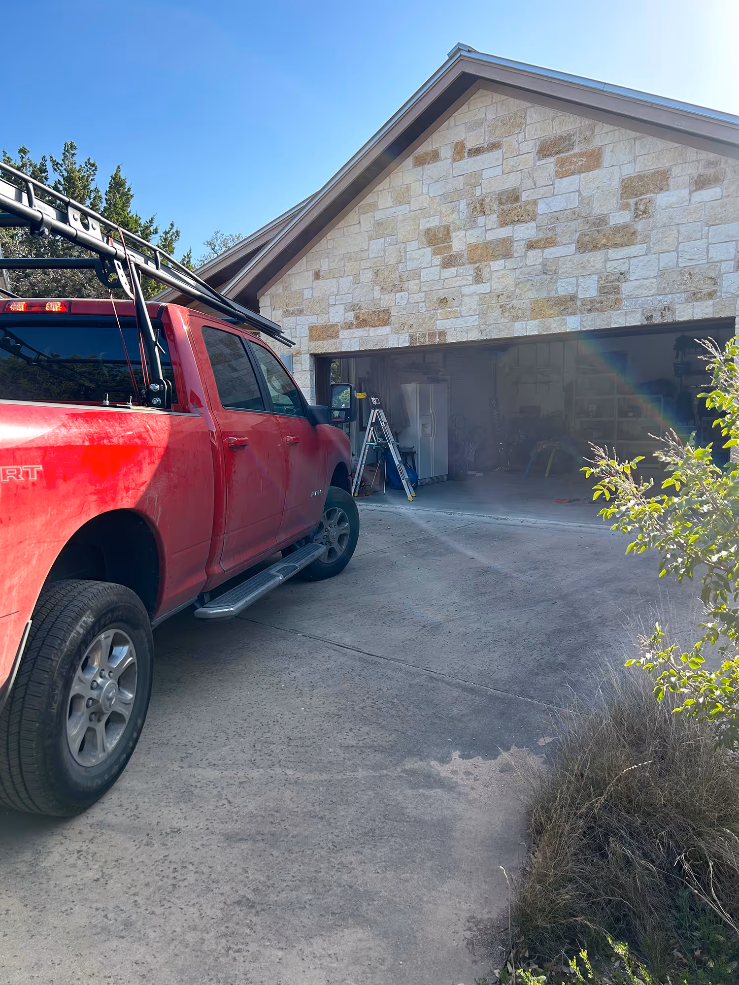 Red pickup truck parked on a driveway next to a stone house with an open garage door showing ladders and tools inside.