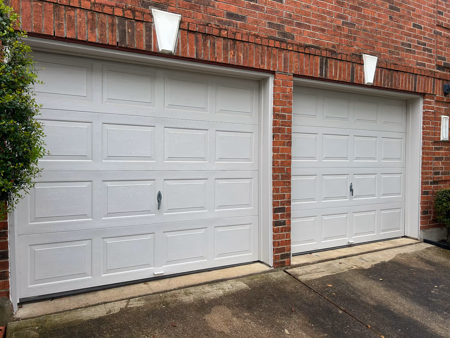 Two closed white garage doors set within a red brick house exterior with concrete driveway in front.