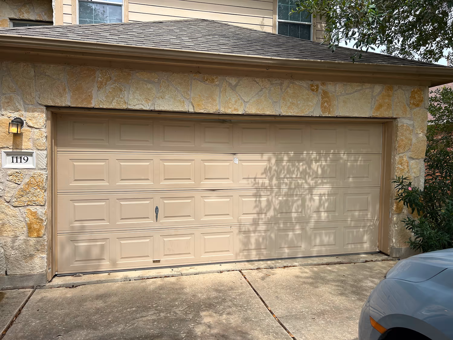 Closed beige double garage door on a stone house with house number 1119 and part of a silver car visible.