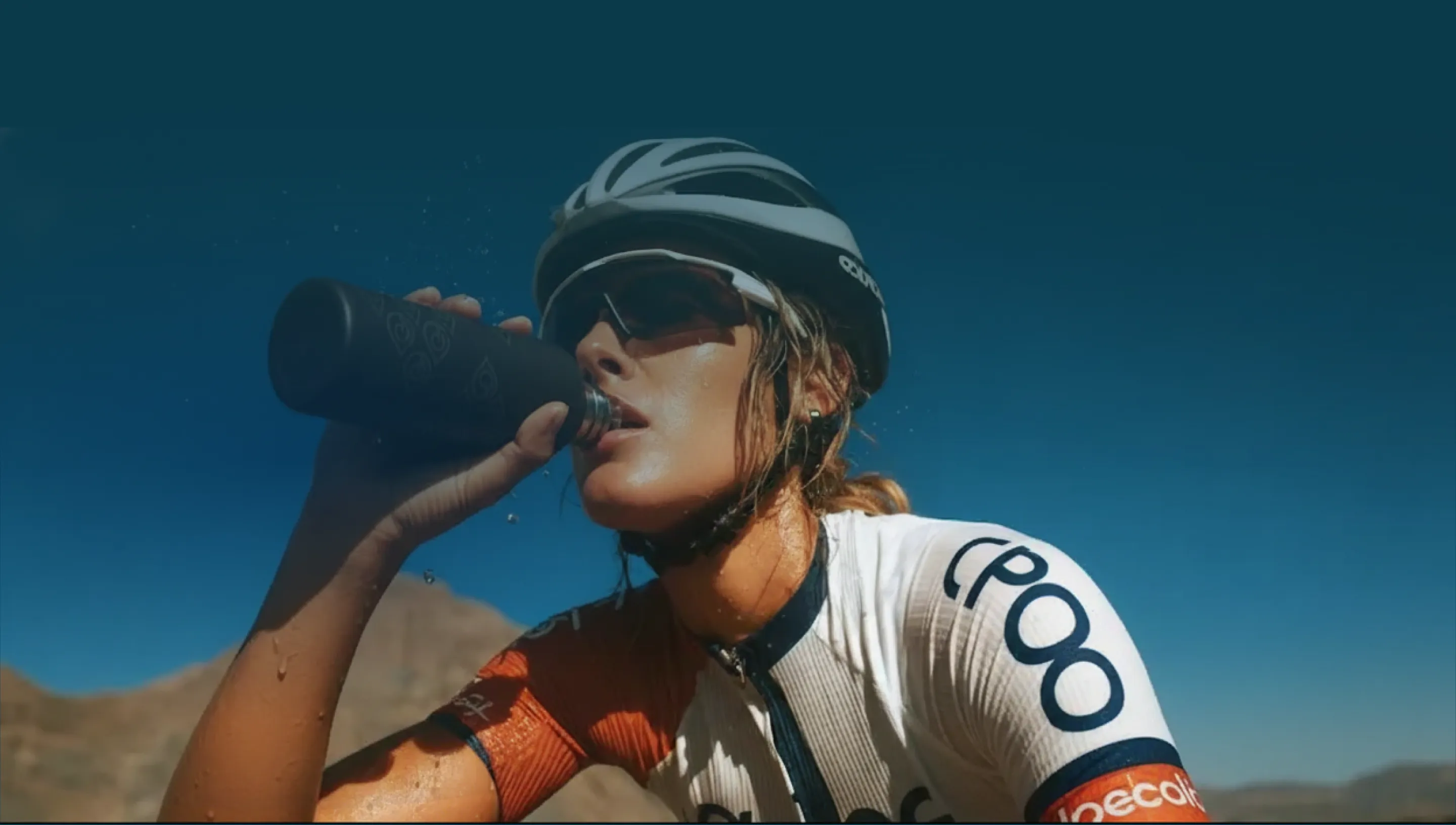 Een fietser met helm en zonnebril die onder een strakblauwe hemel water drinkt uit een fles.