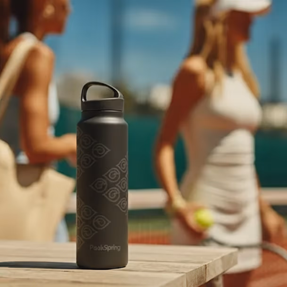 Een zwarte PeakSpring-waterfles met een patroon op een houten tafel, met op de achtergrond twee vrouwen in tenniskleding die wazig zijn.