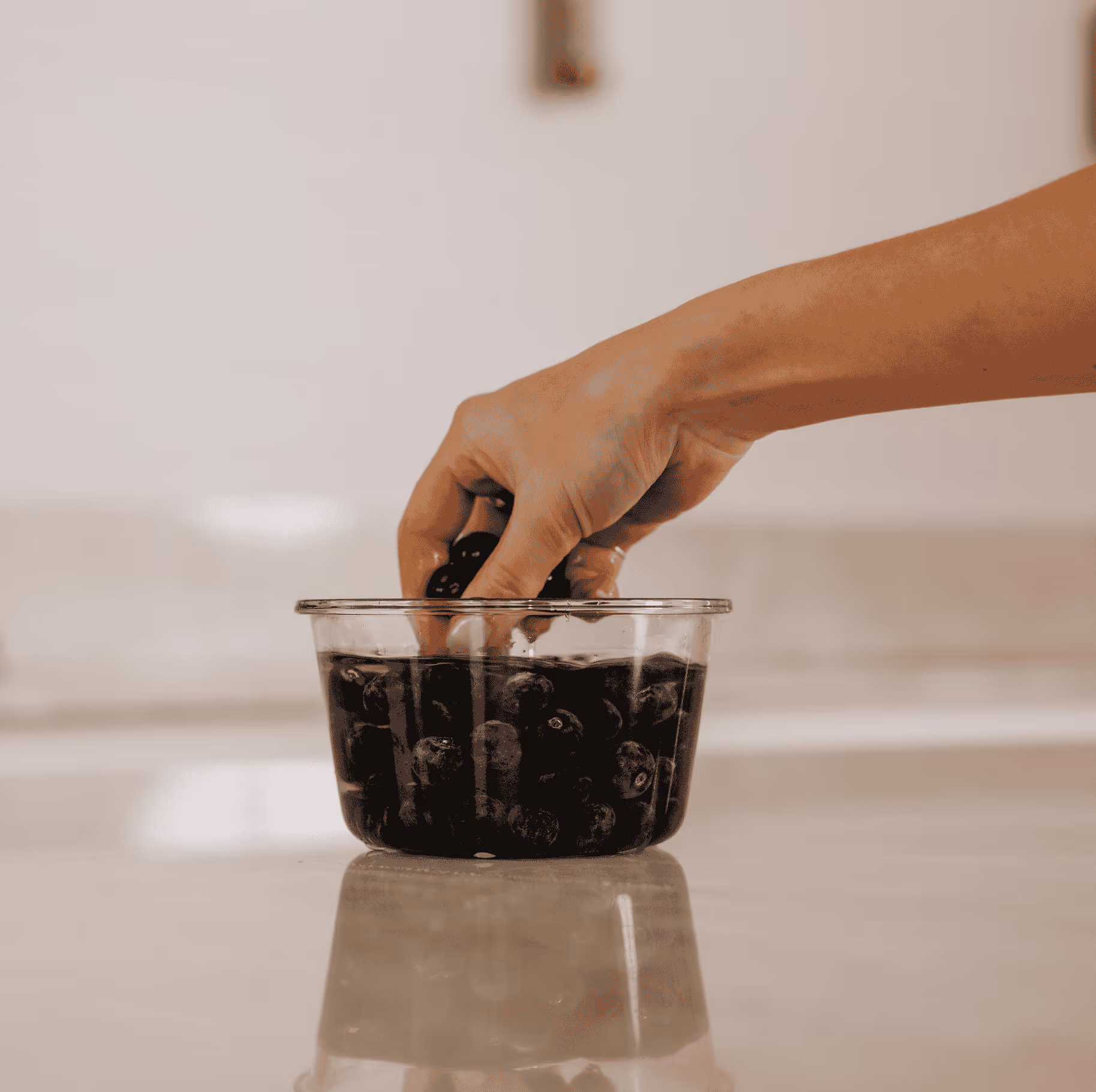 Een hand die in een doorzichtige bak met bosbessen op een spiegelend oppervlak wordt gedompeld.