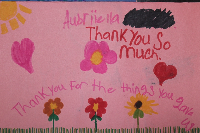 Child's pink thank you card decorated with hand-drawn flowers, hearts, a sun, and handwritten messages saying 'Thank You So Much' and 'Thank you for the things you gave us.' An Artos appreciation note.