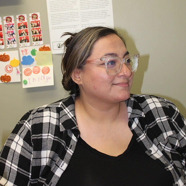 Woman with glasses and a black and white plaid shirt smiling and looking to the side in an office setting. Just one teacher Artos non-profit supports.