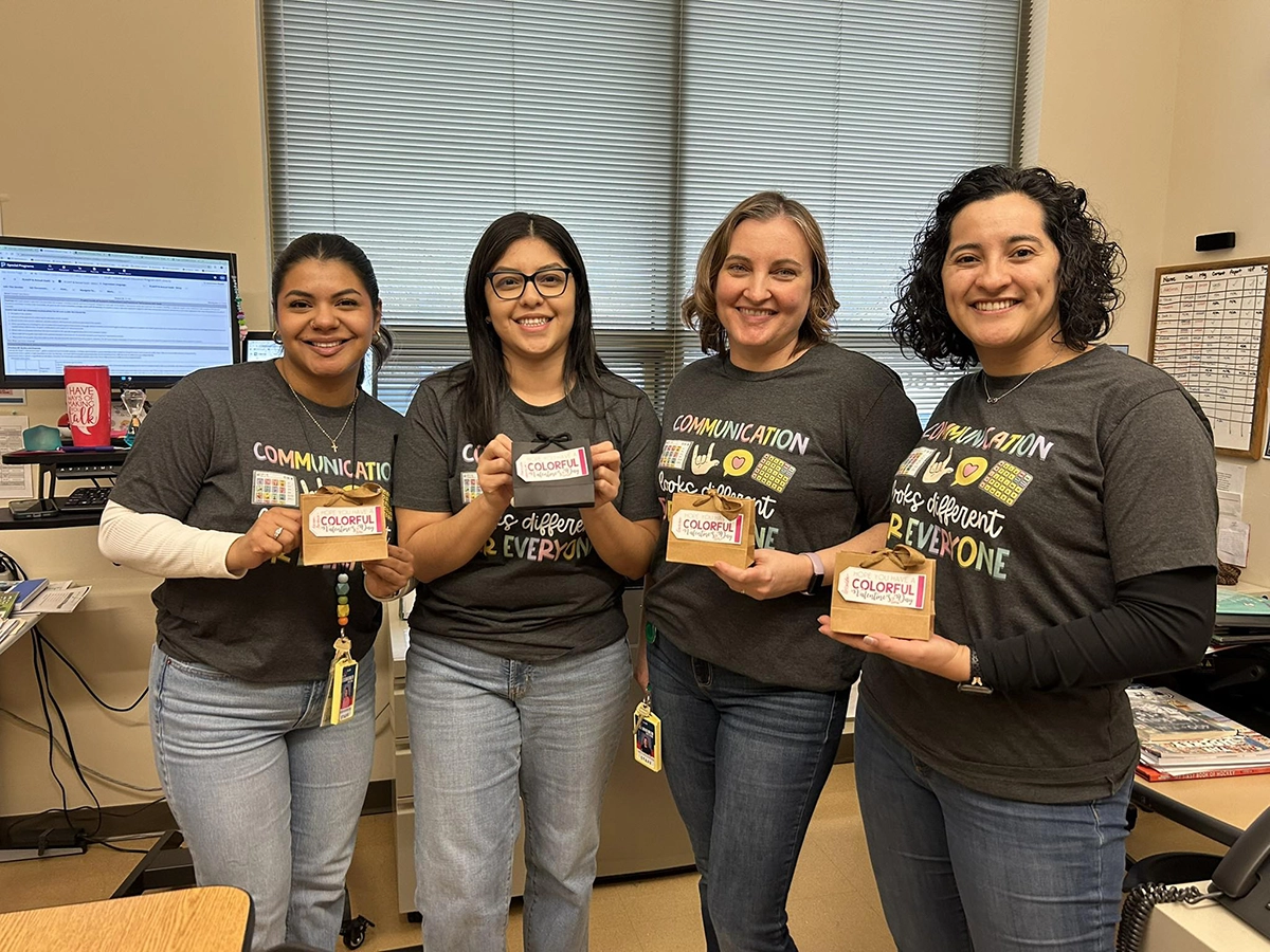 Four women smiling and standing side by side in an office, each holding a small gift box with a colorful Valentine’s Day label, wearing matching dark gray “Communication looks different for everyone” t-shirts.
