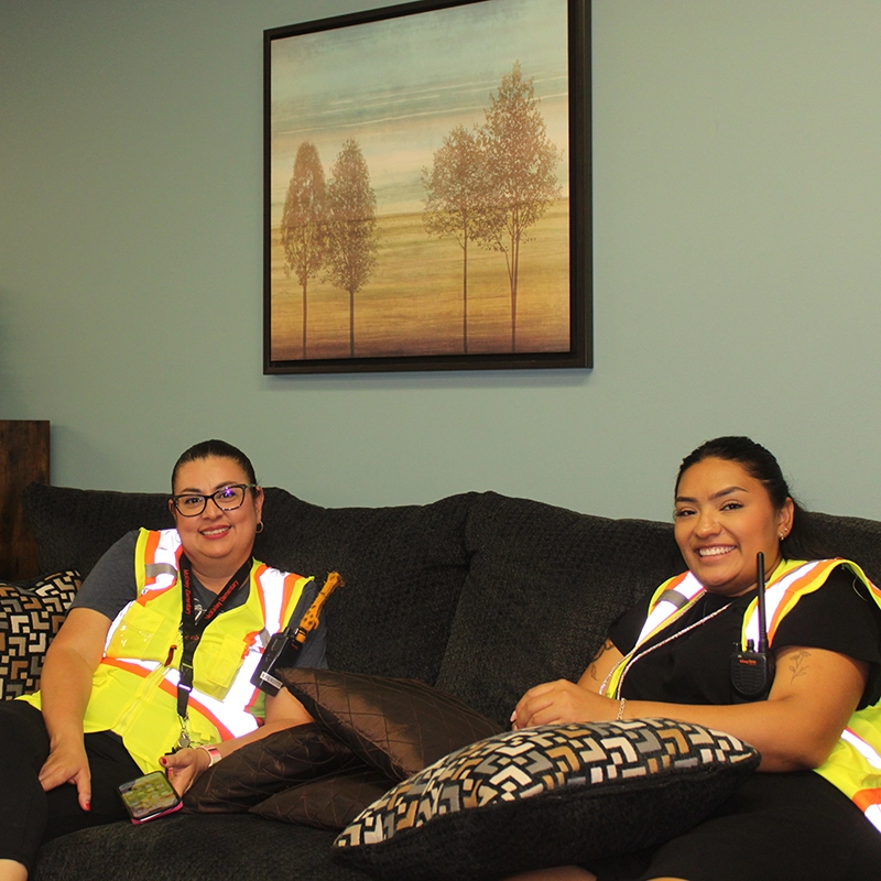 Two women wearing reflective safety vests sitting and smiling on a gray couch with patterned and solid pillows, under a framed painting of trees in the new lounge provided by Artos Yakima Non-profit.