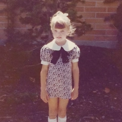 Young girl Amy with blonde hair tied up, wearing a short patterned dress with a large white collar and black bow, standing outdoors in front of a brick wall.