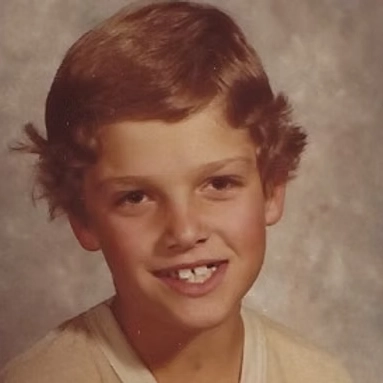 Portrait of a smiling young Russ with short brown hair and missing front teeth against a neutral background.